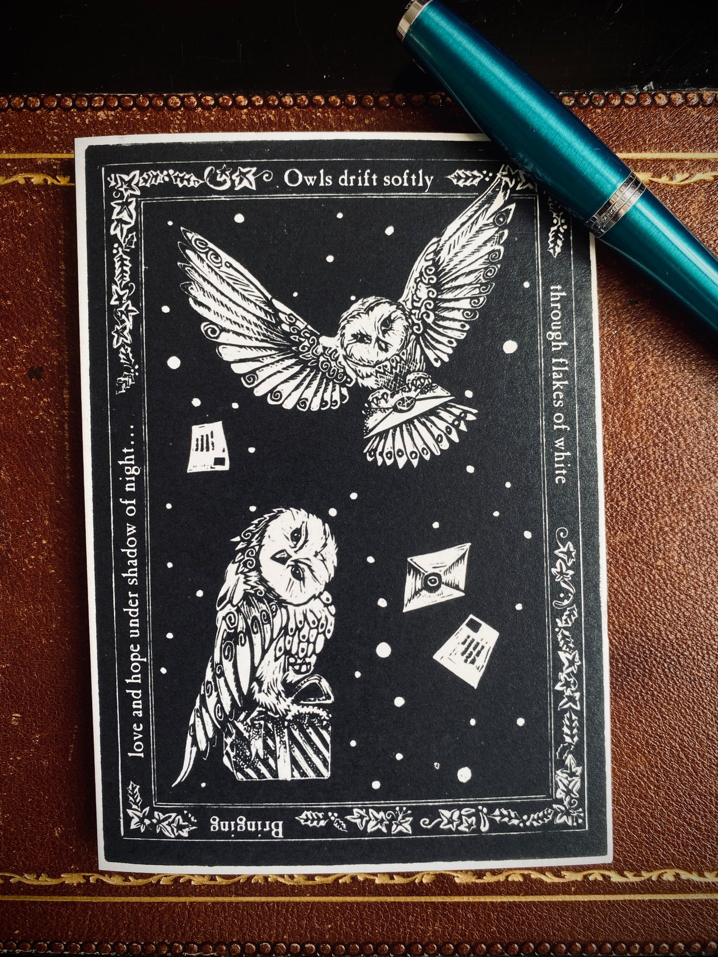 Black and white owl illustration on a card with decorative elements, placed on a brown leather surface with a pen.