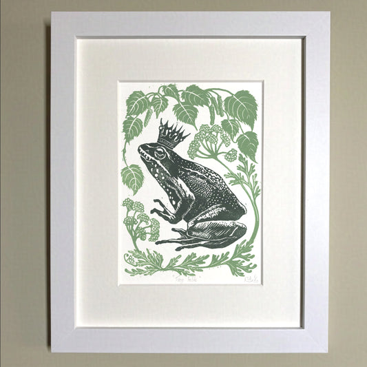 Framed artwork of a frog with a crown surrounded by green leaves on a beige wall.