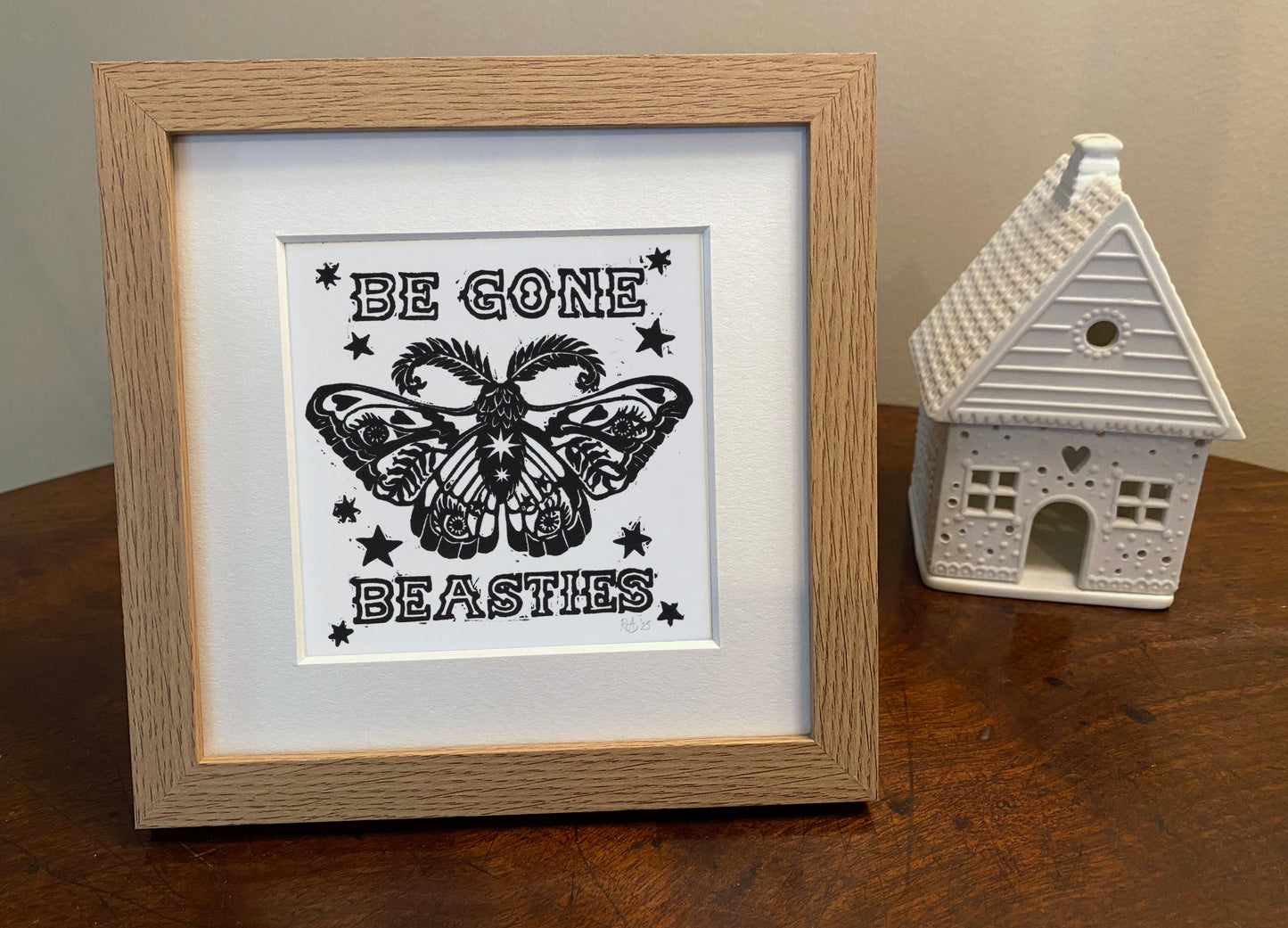 Framed artwork with moth design and text 'Be Gone Beasties' on a wooden surface.