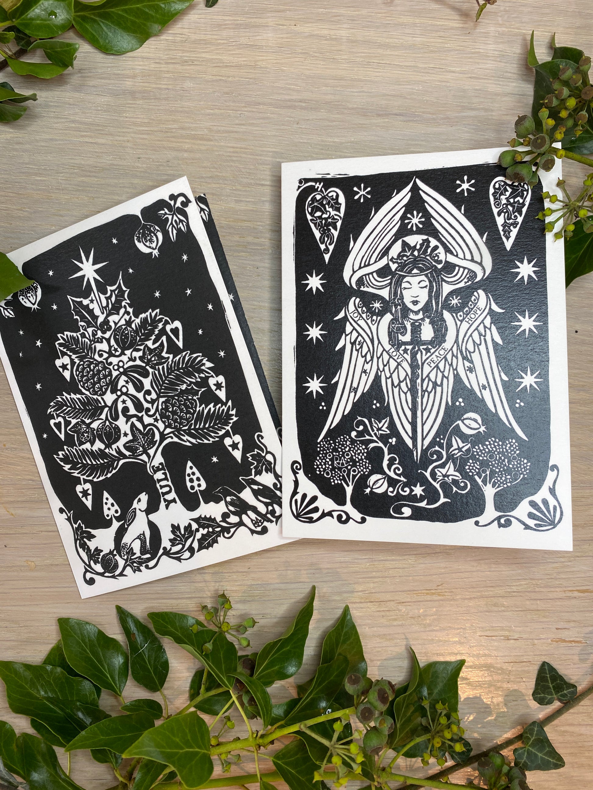 Two black and white christmas cards on a wooden surface with green leaves.
