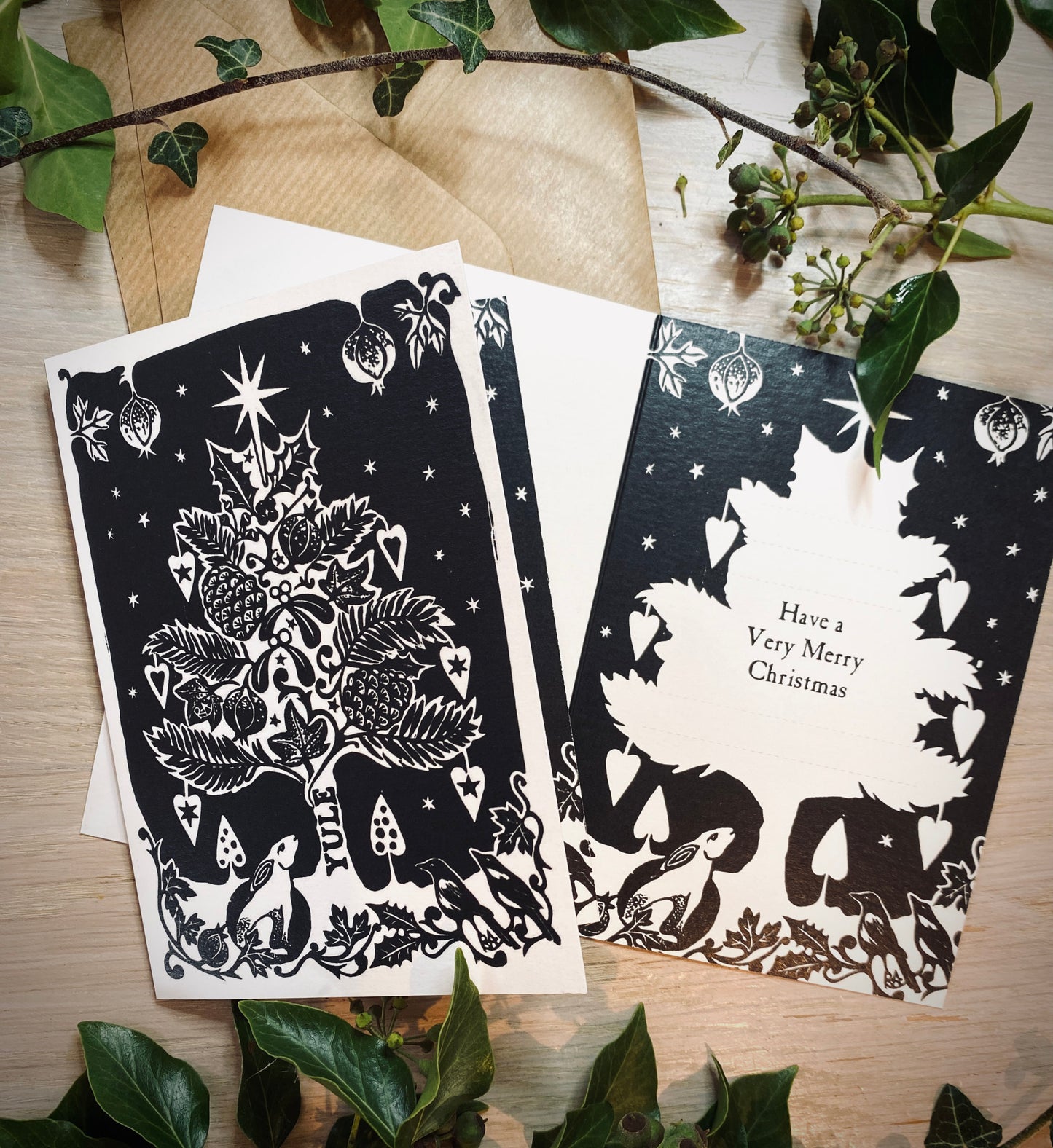Two black and white Christmas-themed cards on a wooden surface with greenery.