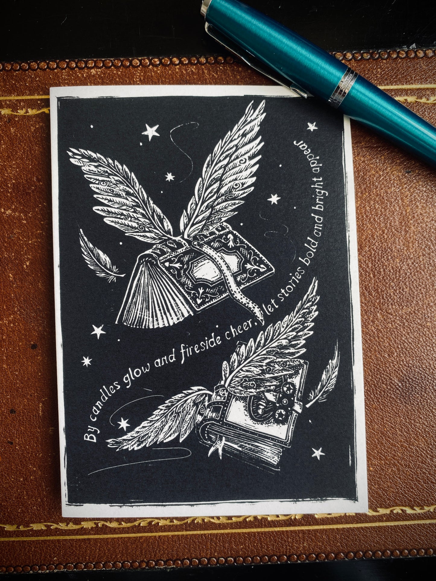 Decorative card with winged book design on a leather surface with a pen.