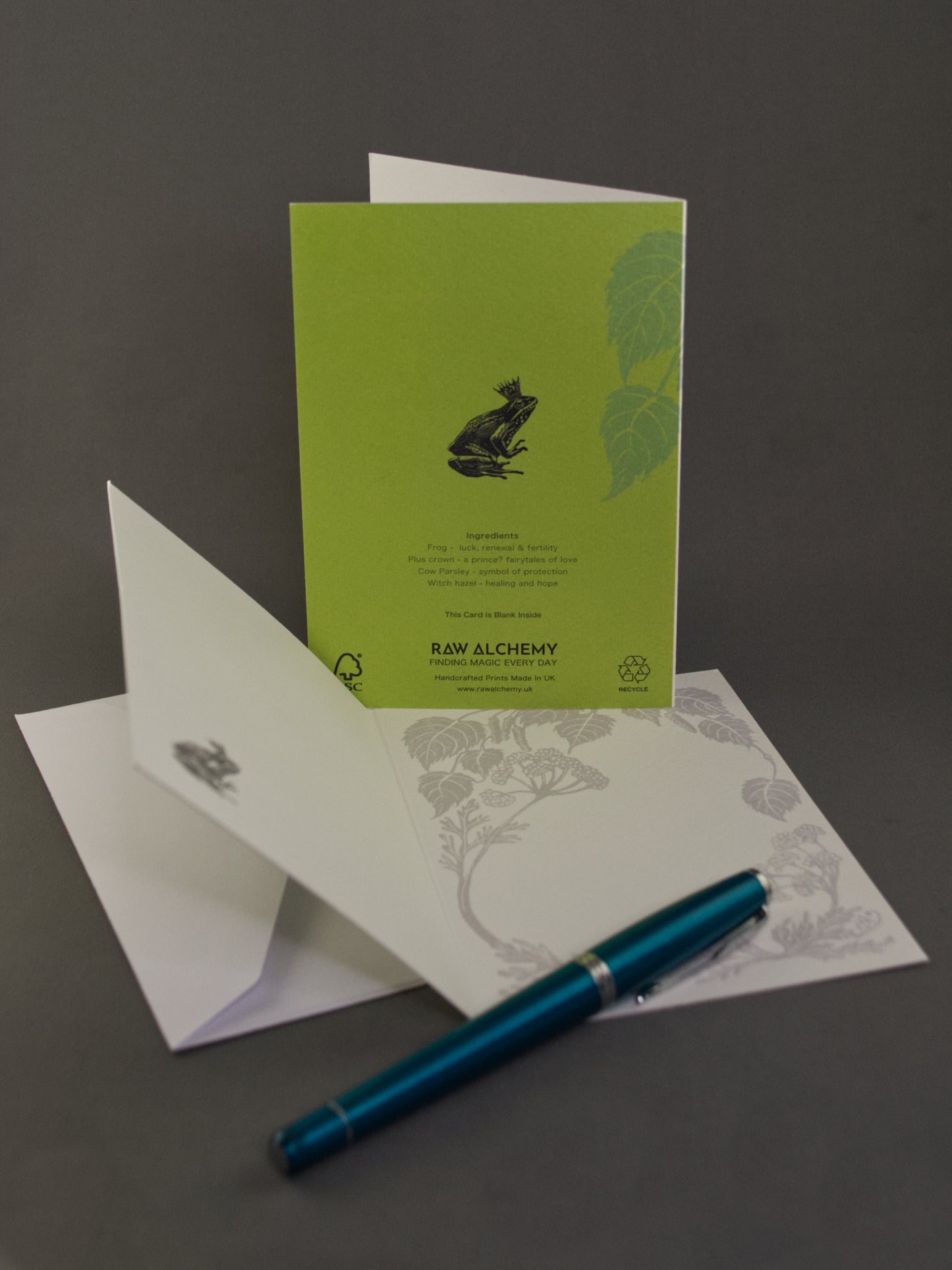 Green greeting card with a frog design, white envelope, and blue pen on a gray background