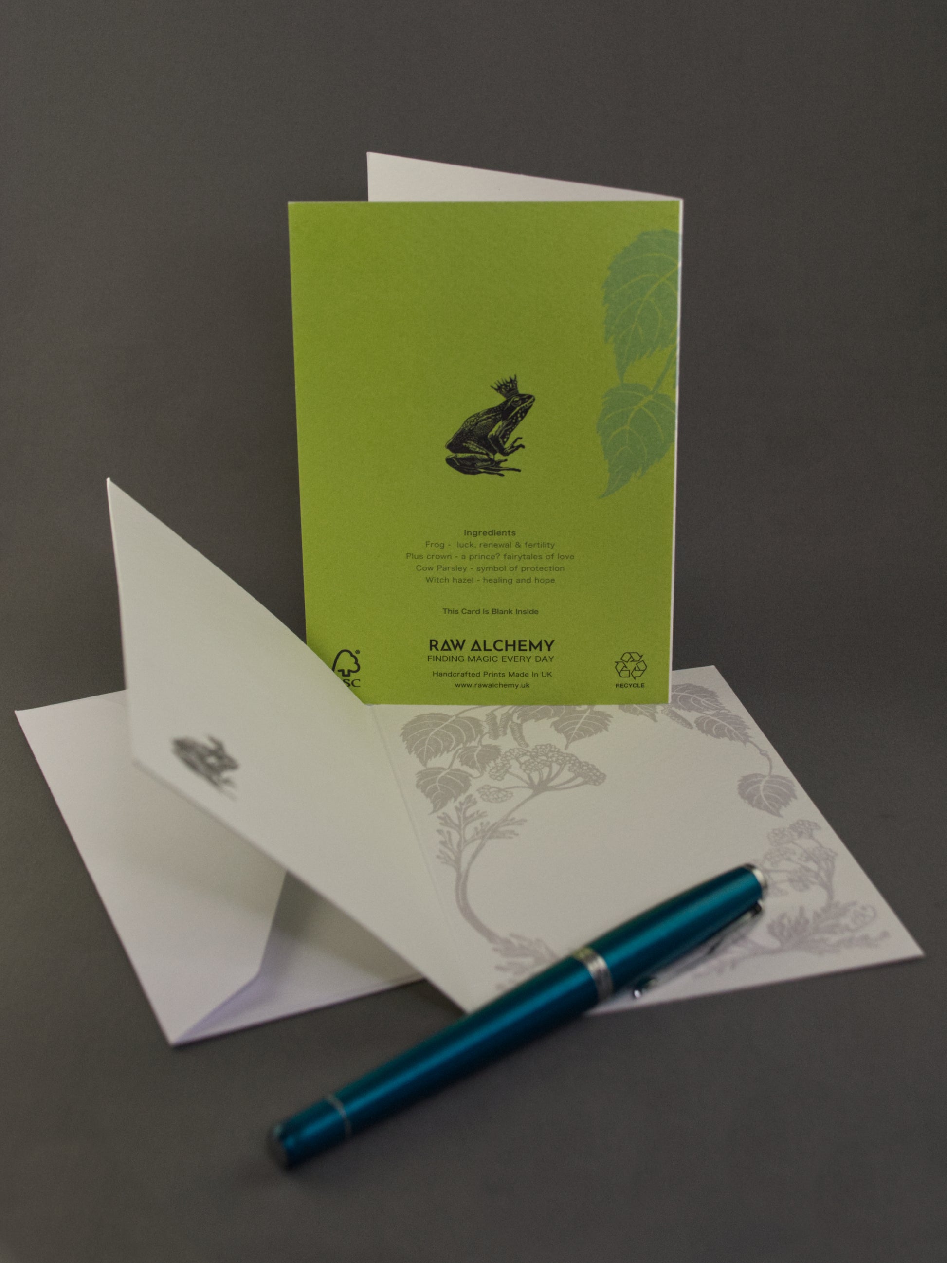 Green greeting card with a frog design, white envelope, and blue pen on a gray background