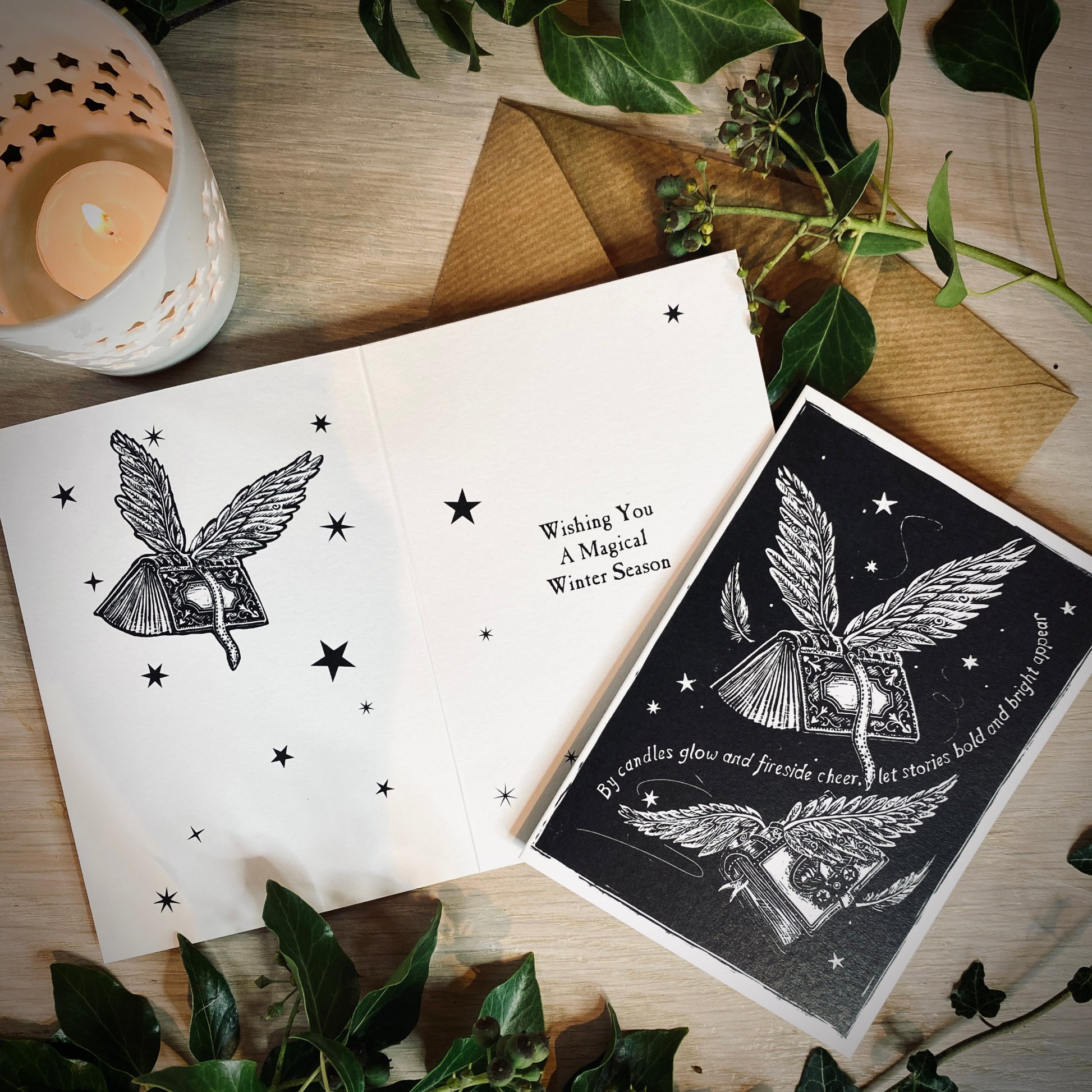 Two greeting cards with winged heart designs on a wooden surface with leaves and a candle.