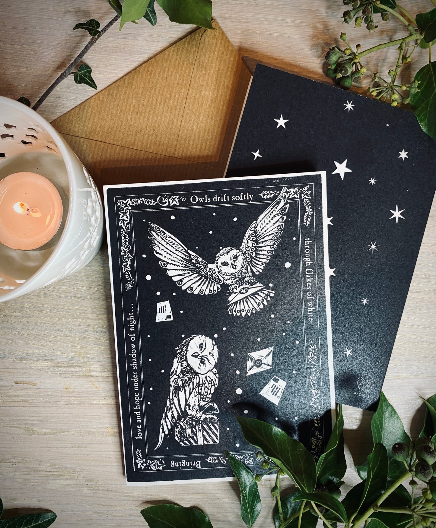 Black card with owl design on a wooden surface with a candle and plants.