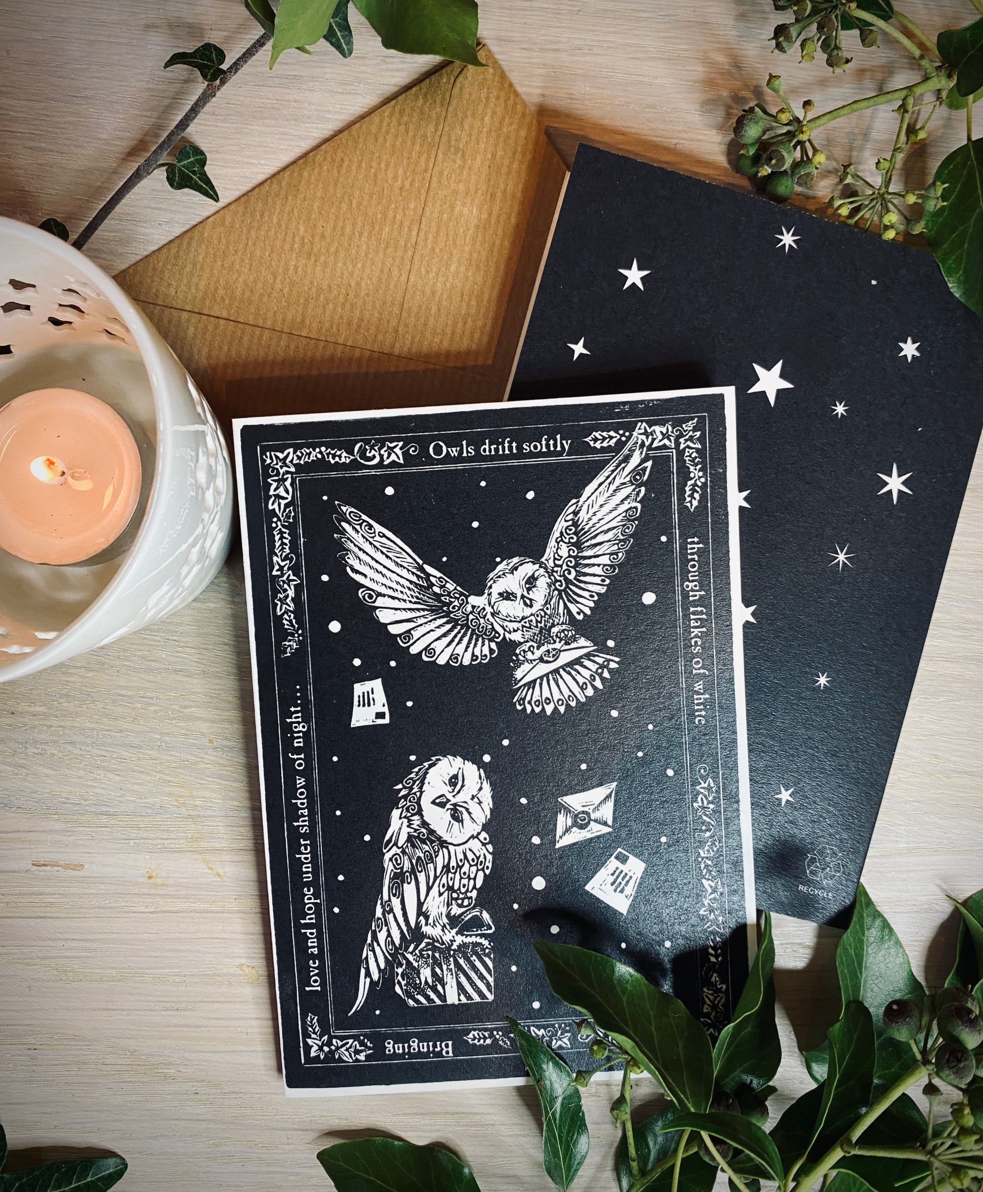 Black card with owl design on a wooden surface with a candle and plants.