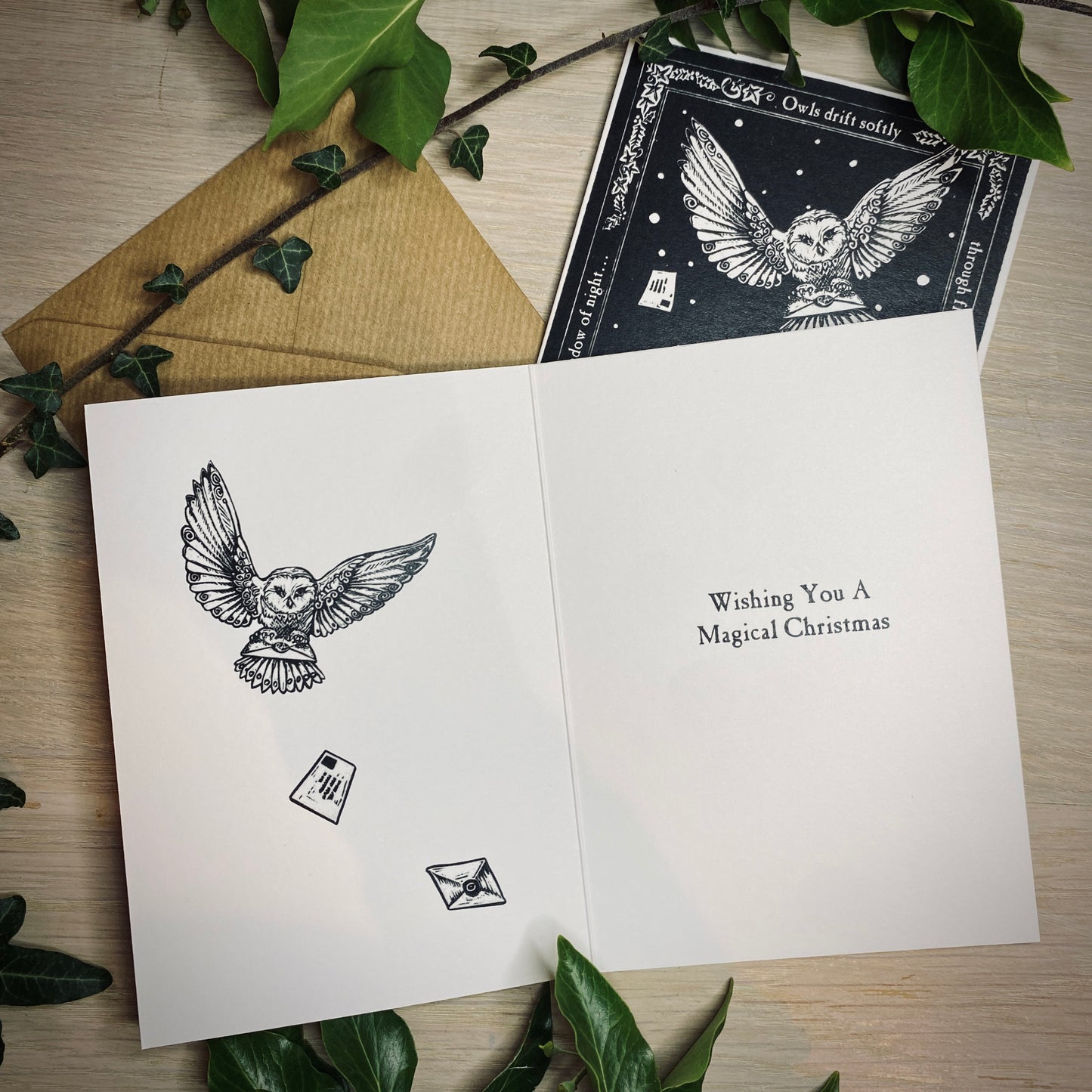 Open greeting card with owl illustration and 'Wishing You A Magical Christmas' text, surrounded by green leaves on a wooden surface.