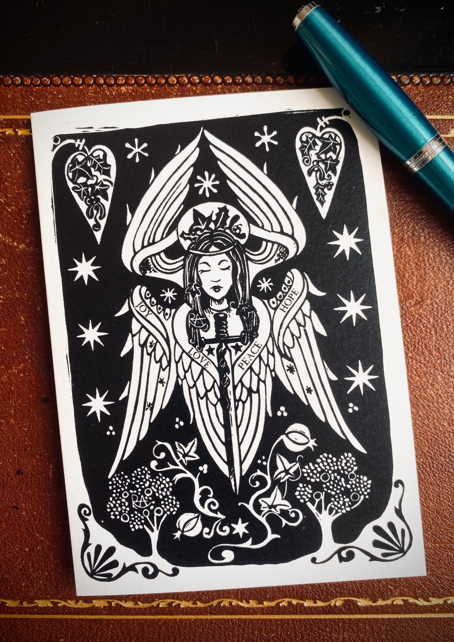 Black and white angel illustration on a piece of paper with a pen on a brown leather surface.