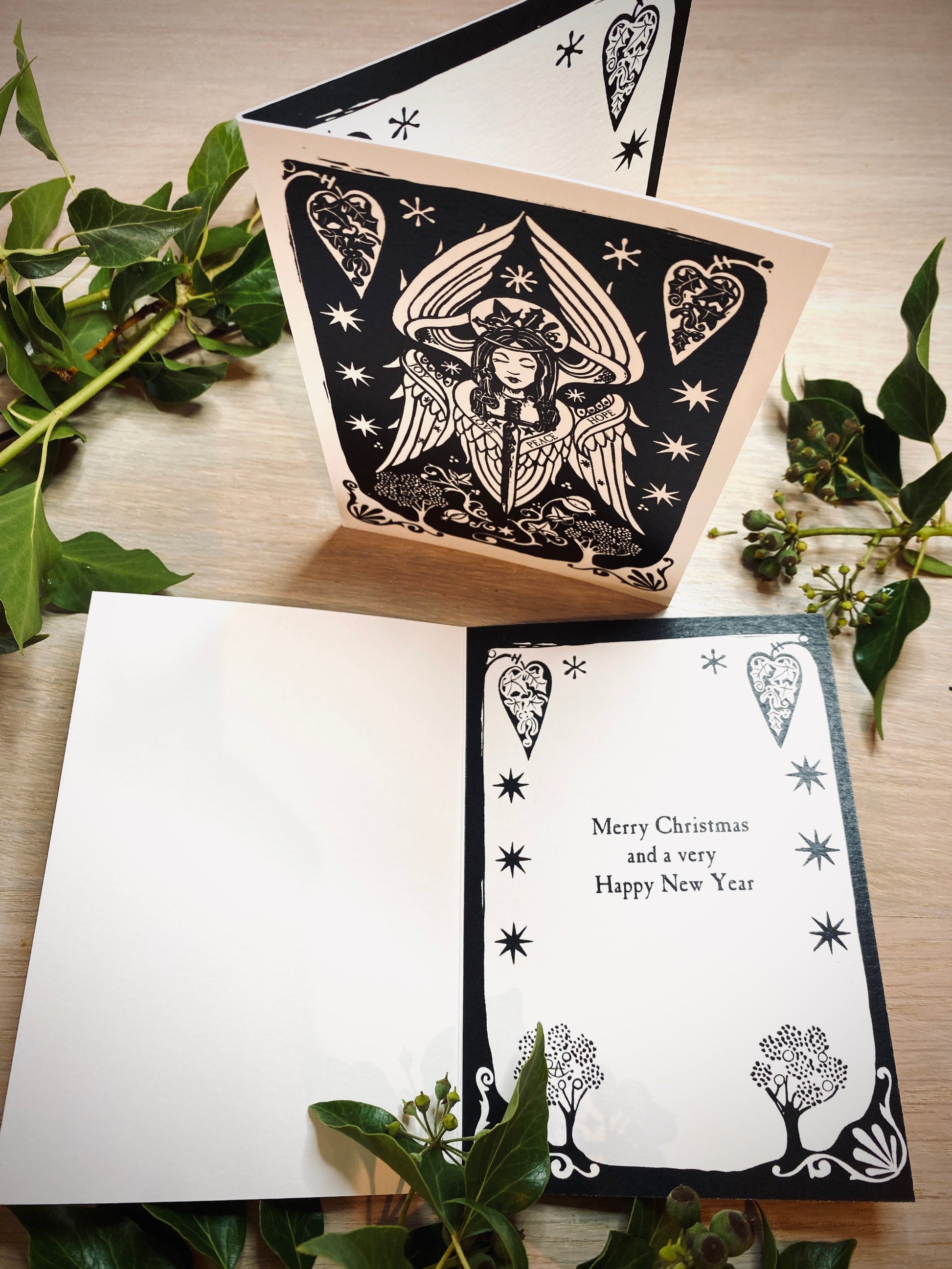 Black and white angel greeting cards with decorative designs on a wooden surface with greenery.