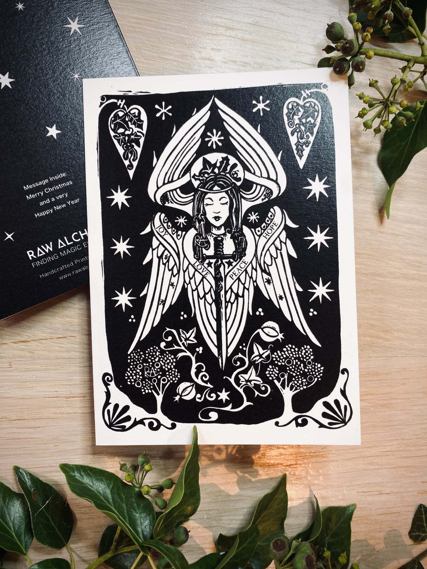 Black and white illustration of a winged figure on a white card with decorative elements, placed on a wooden surface with green leaves.