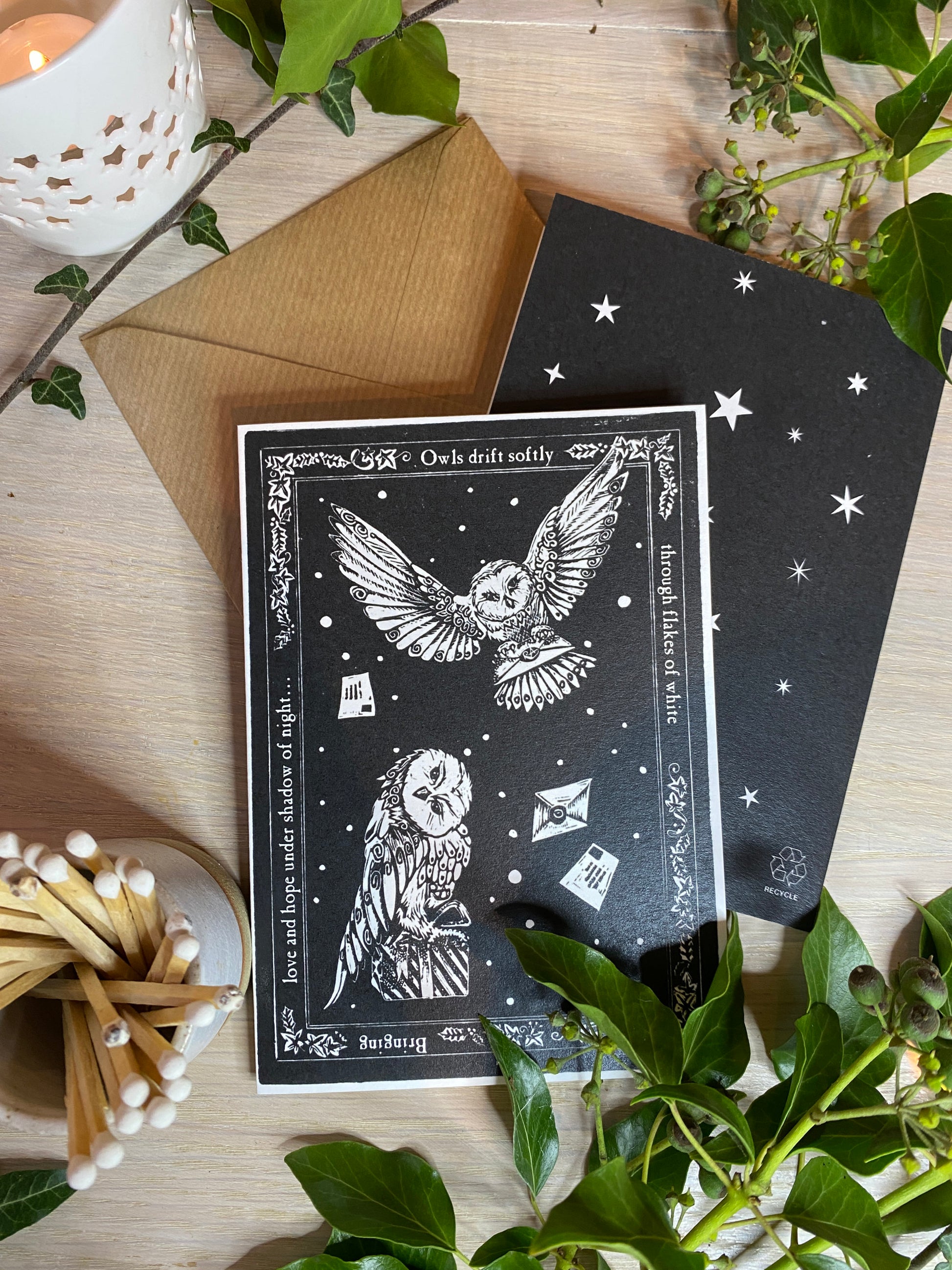 Black card with owl design on a wooden surface with plants and matches.