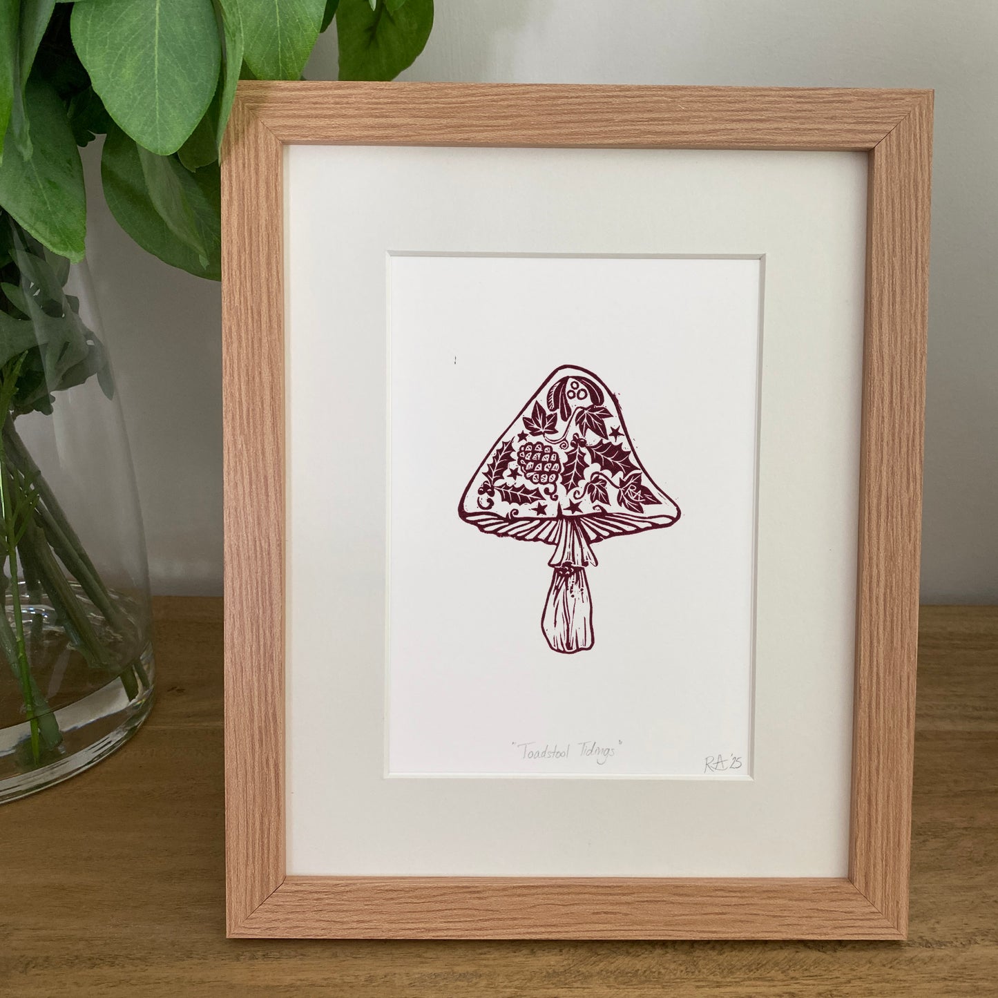 Framed illustration of a decorative mushroom on a wooden surface with a plant in the background.