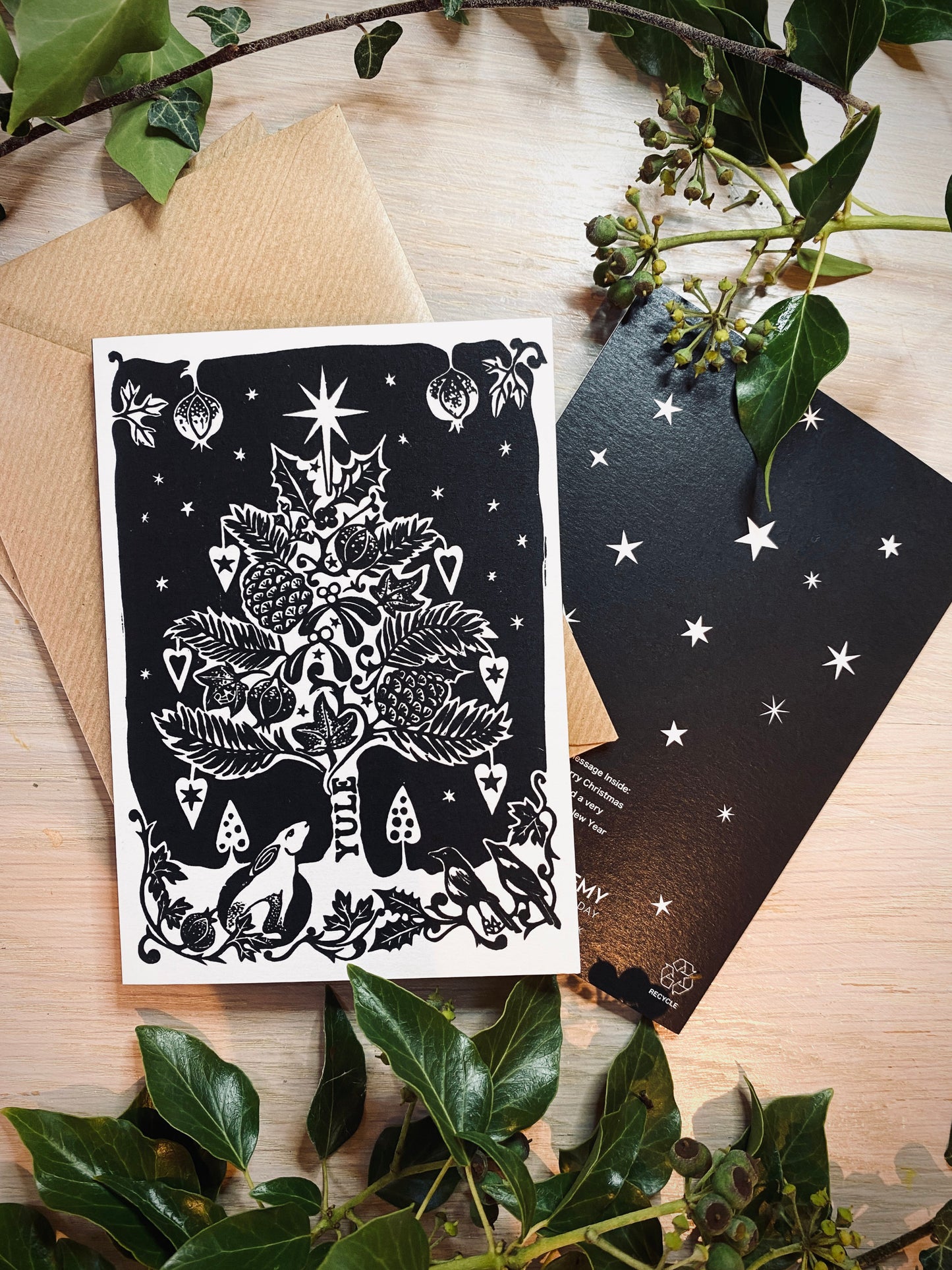 Black and white Christmas-themed card with tree and hare design on a wooden surface with greenery.