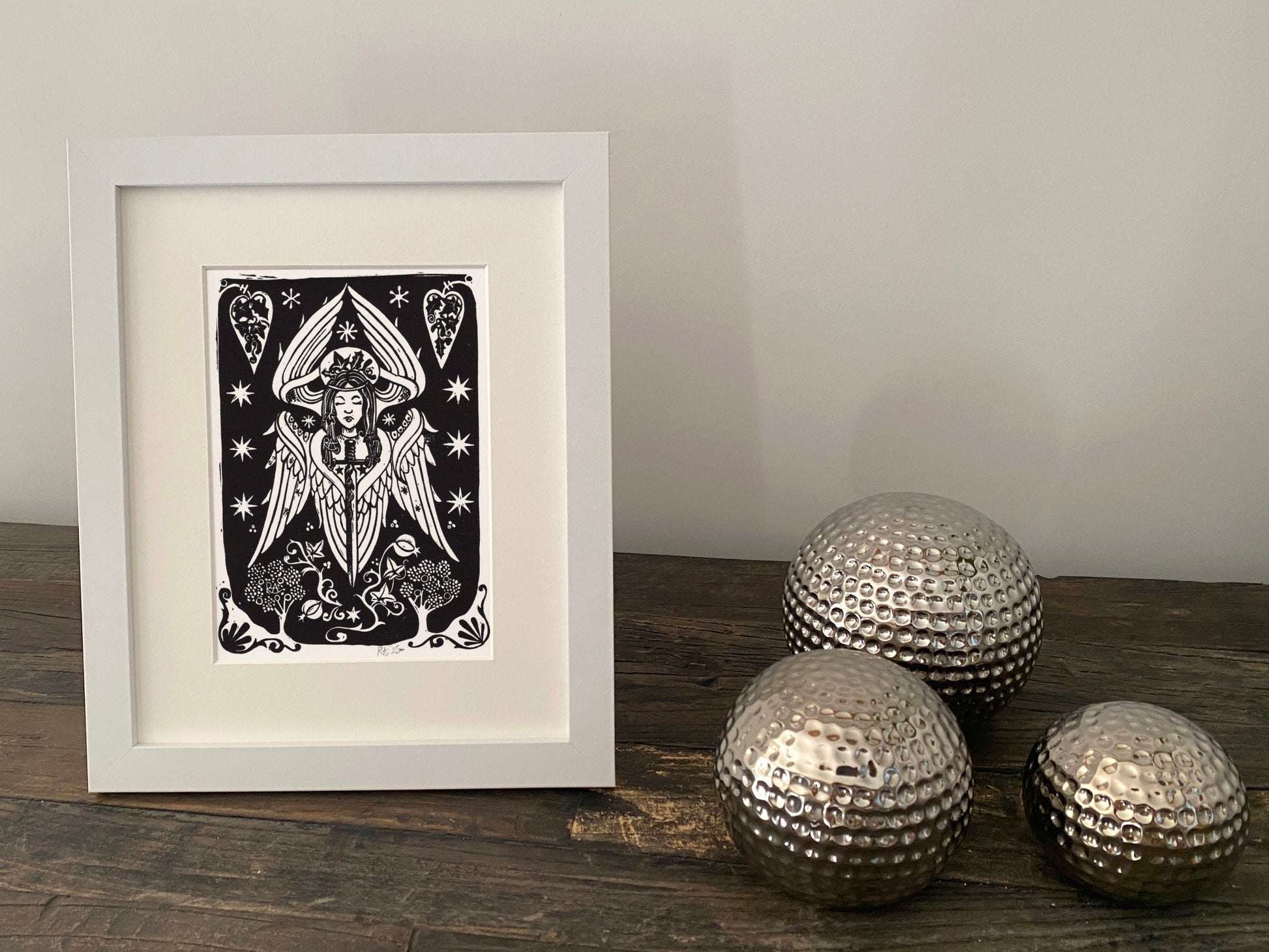 Framed artwork of a black and white angel design on a wooden surface with decorative silver balls.