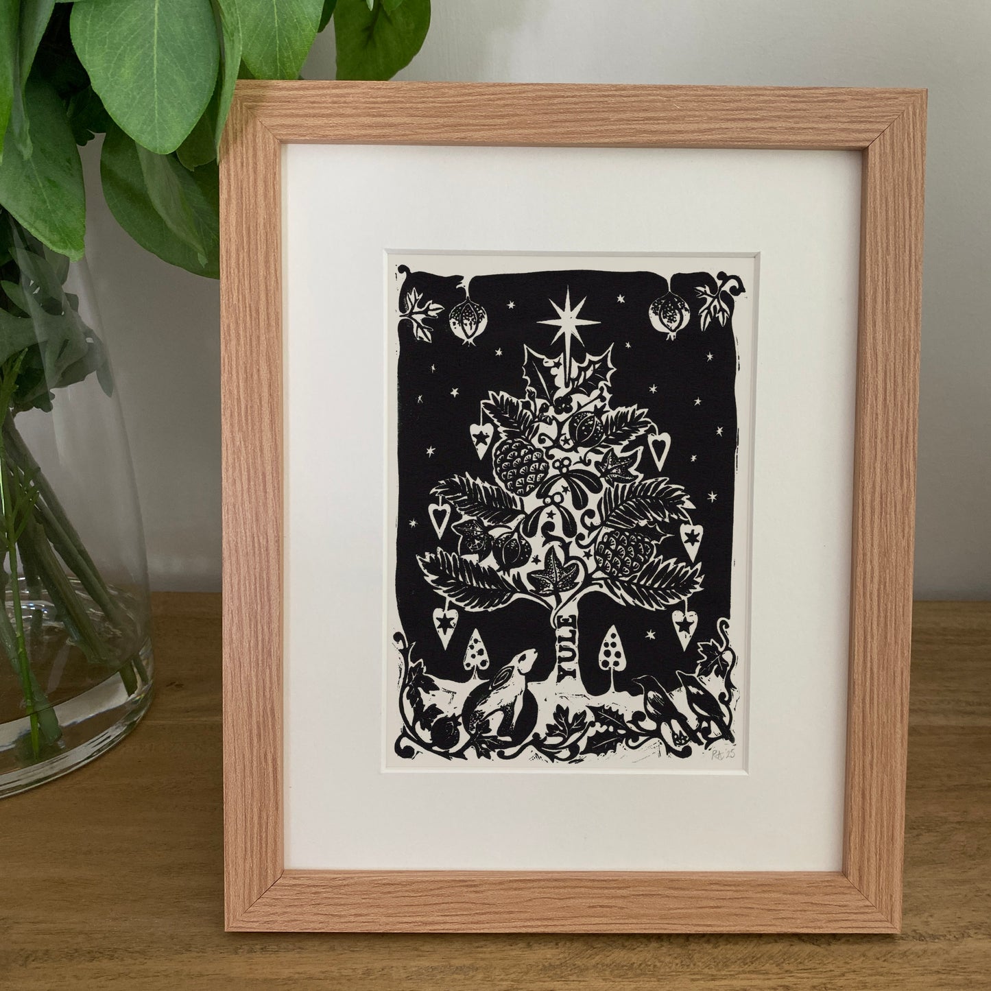 Framed black and white artwork of a Christmas tree on a wooden surface with a plant in the background.