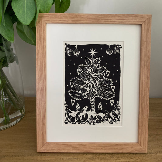 Framed black and white artwork of a Christmas tree on a wooden surface with a plant in the background.