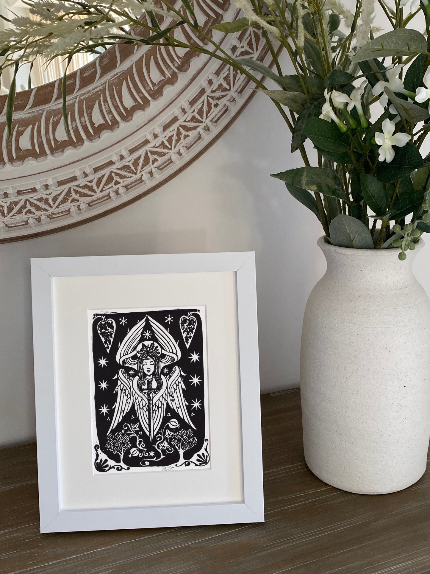 Framed black and white angel artwork on a wooden surface with a vase of flowers in the background.