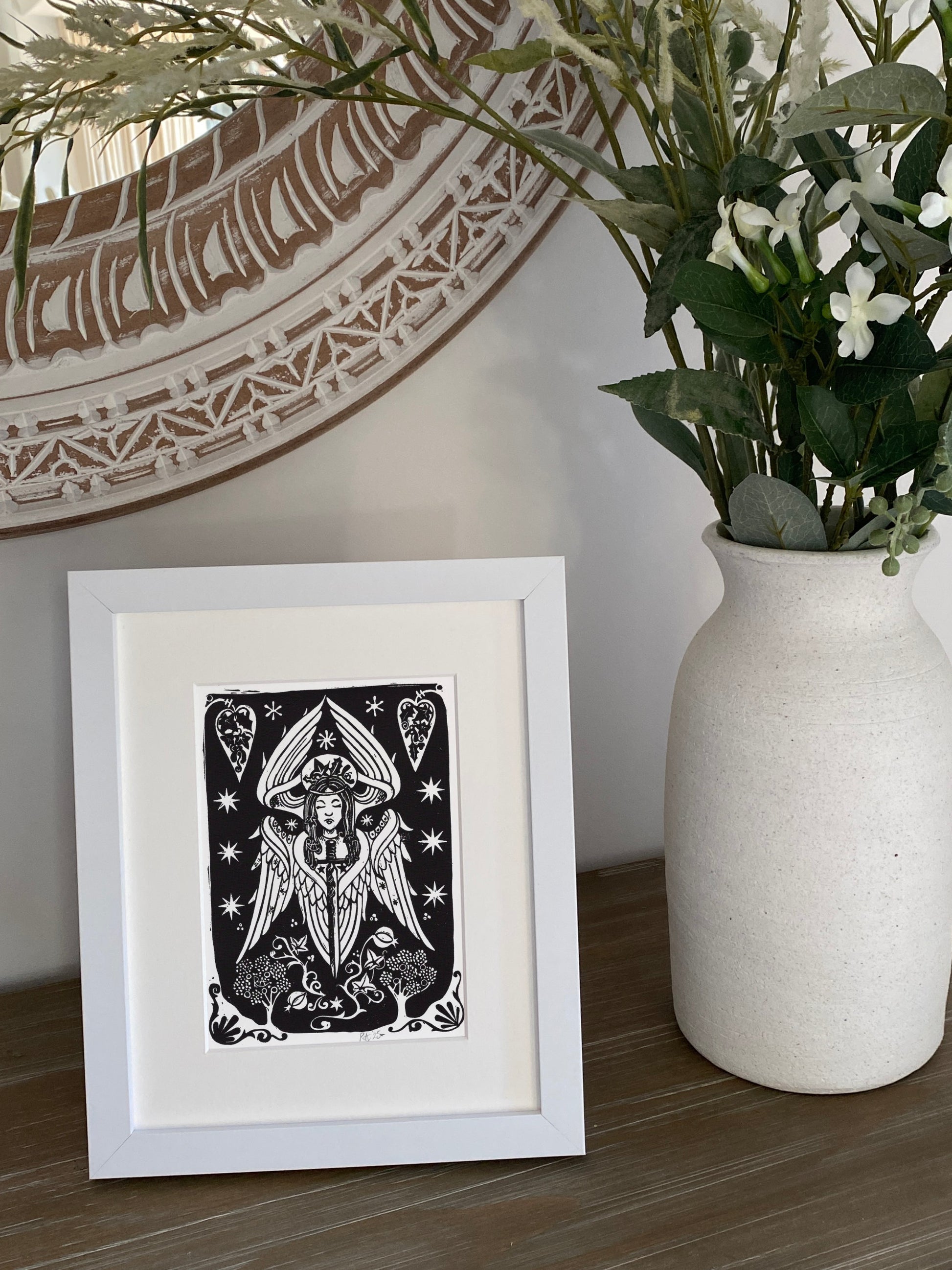 Framed black and white angel artwork on a wooden surface with a vase of flowers in the background.