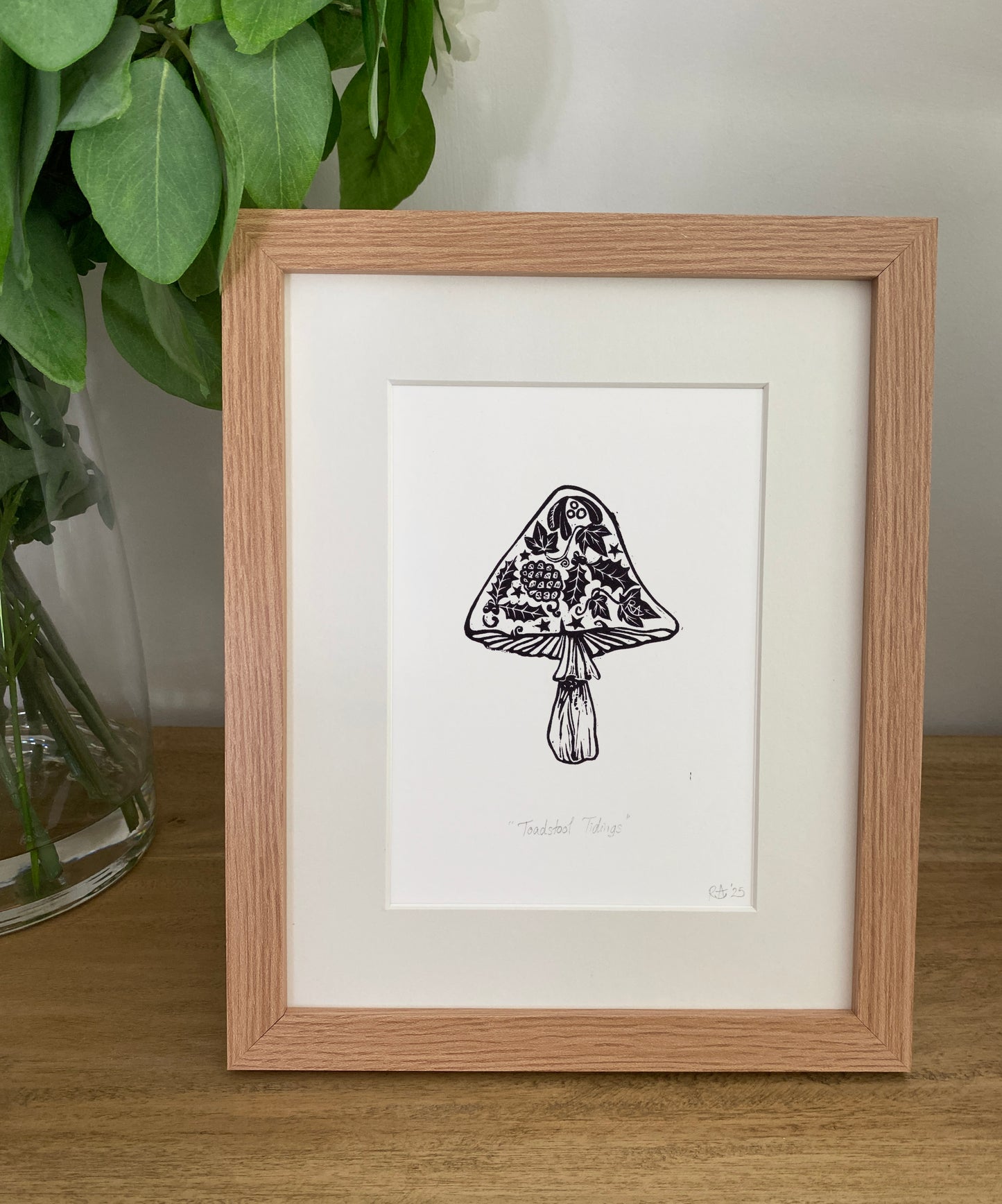 Framed black and white illustration of a mushroom on a wooden surface with greenery in the background.