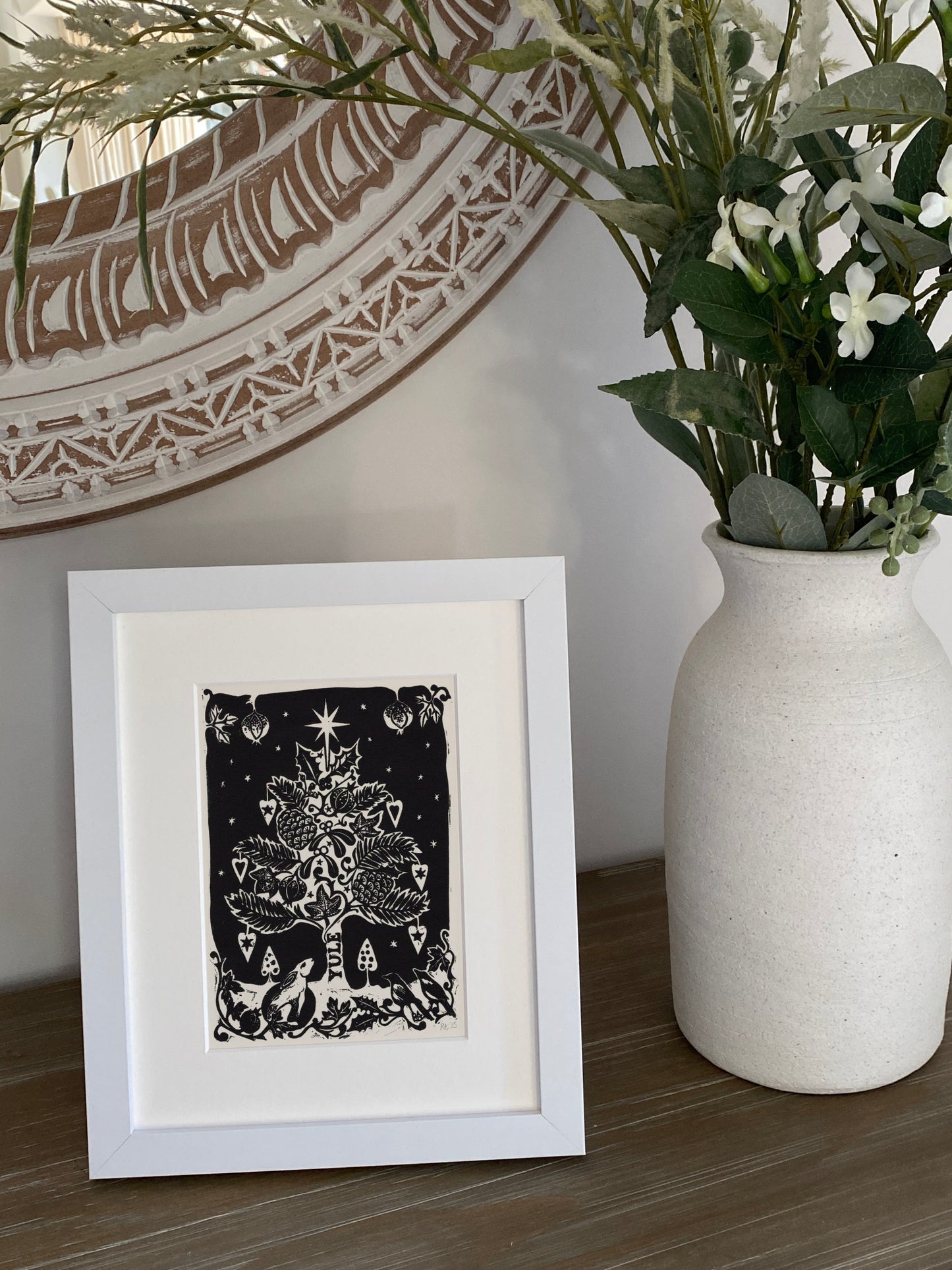 Framed artwork next to a vase with plants on a wooden surface