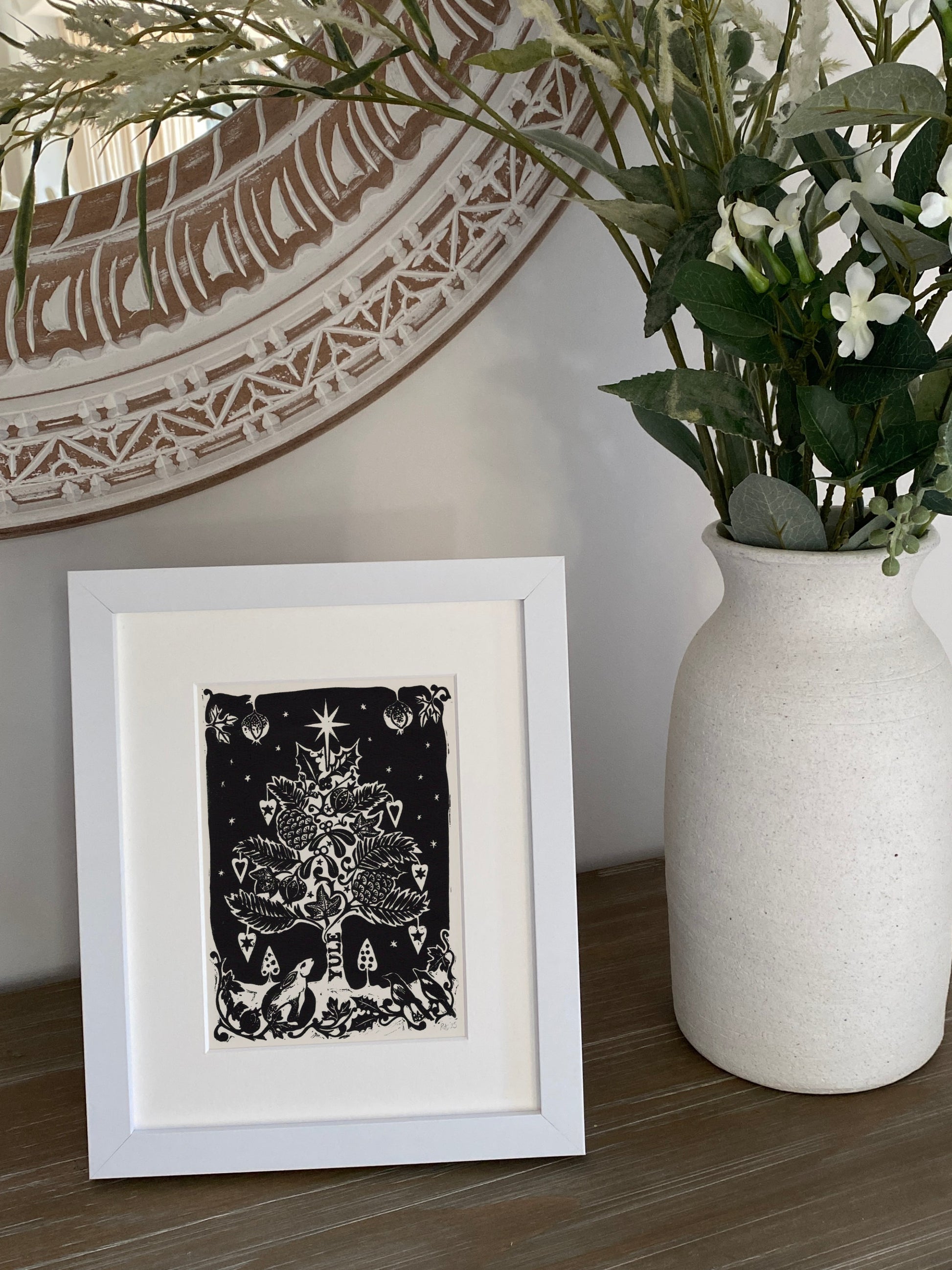 Framed artwork next to a vase with plants on a wooden surface