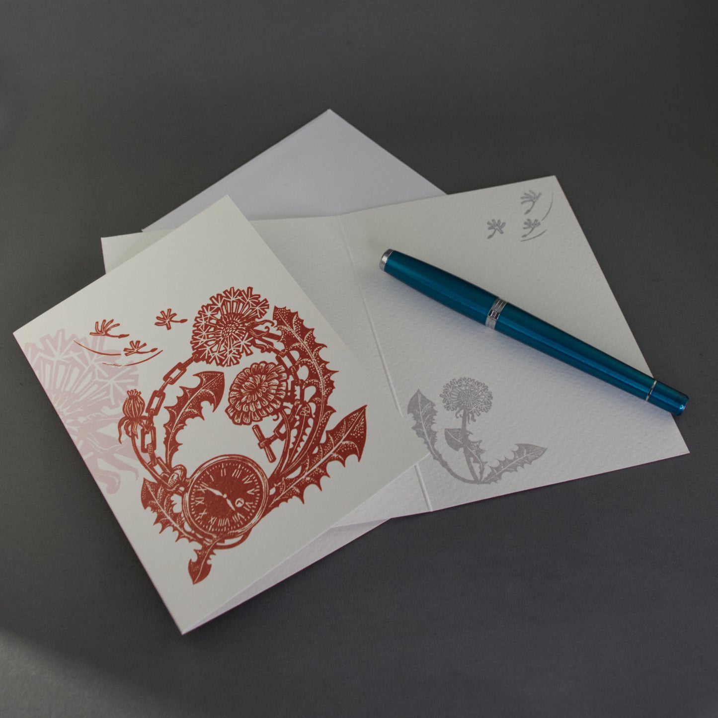 Greeting card with dandelion floral design, pen, and envelope on a dark surface