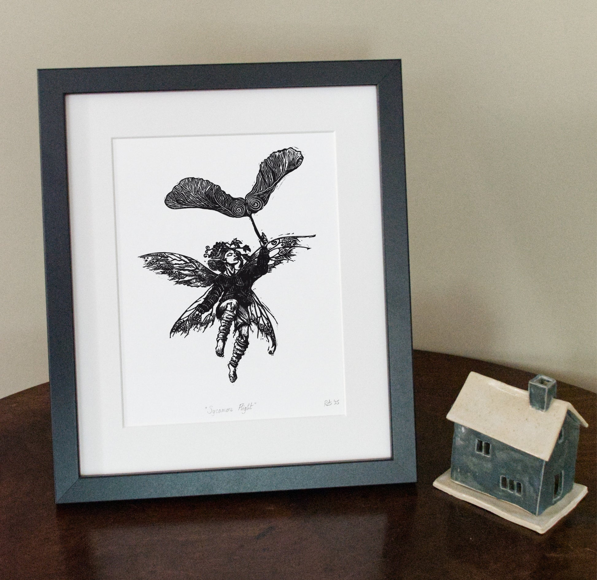 Framed black and white illustration of a fairy on a white background, next to a small model house.