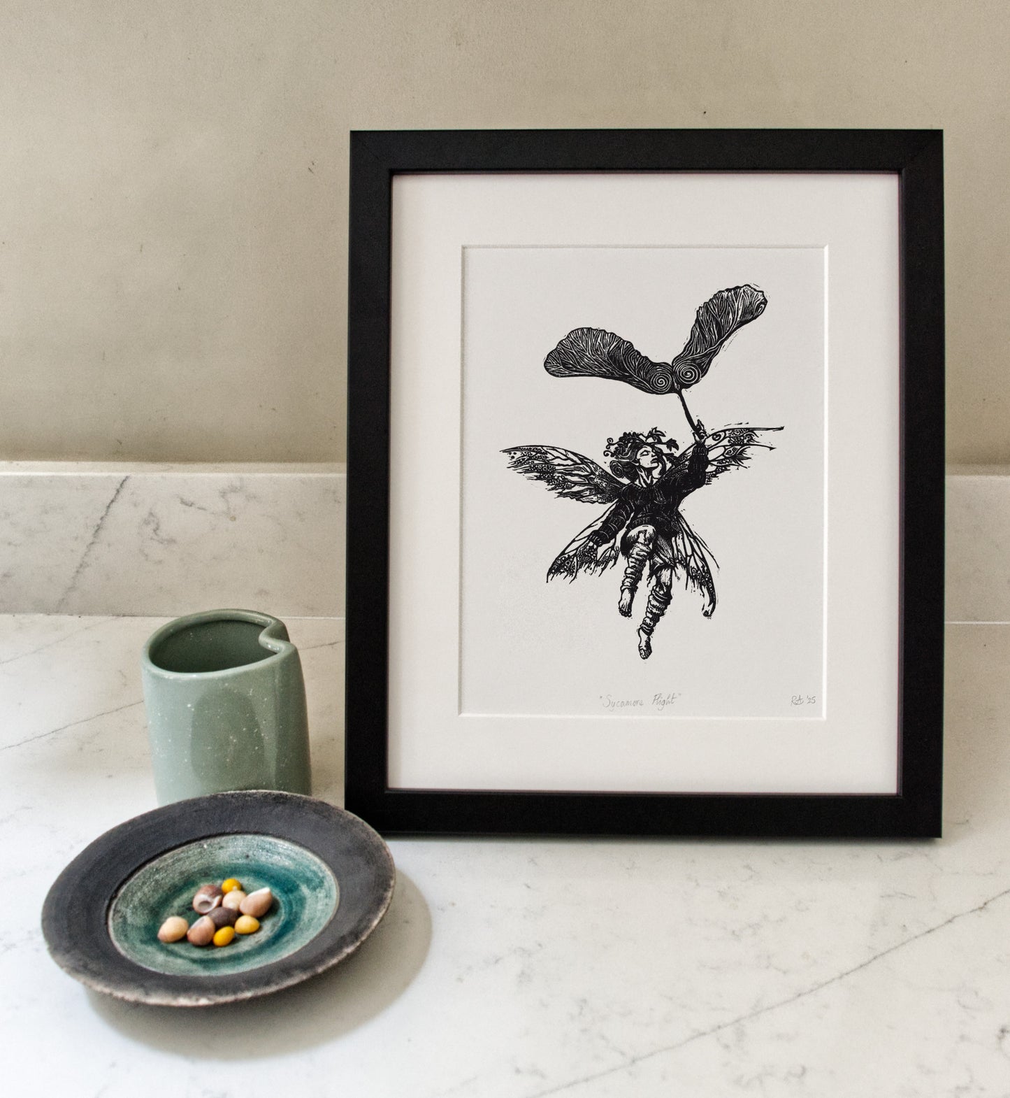 Framed black and white artwork of a fairy with a leaf on a white surface.