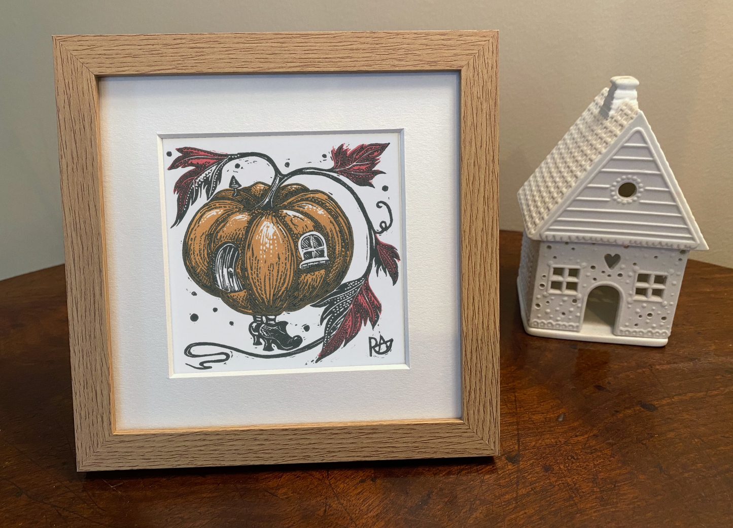 Framed artwork of a pumpkin with decorative elements on a wooden surface next to a small house-shaped object.