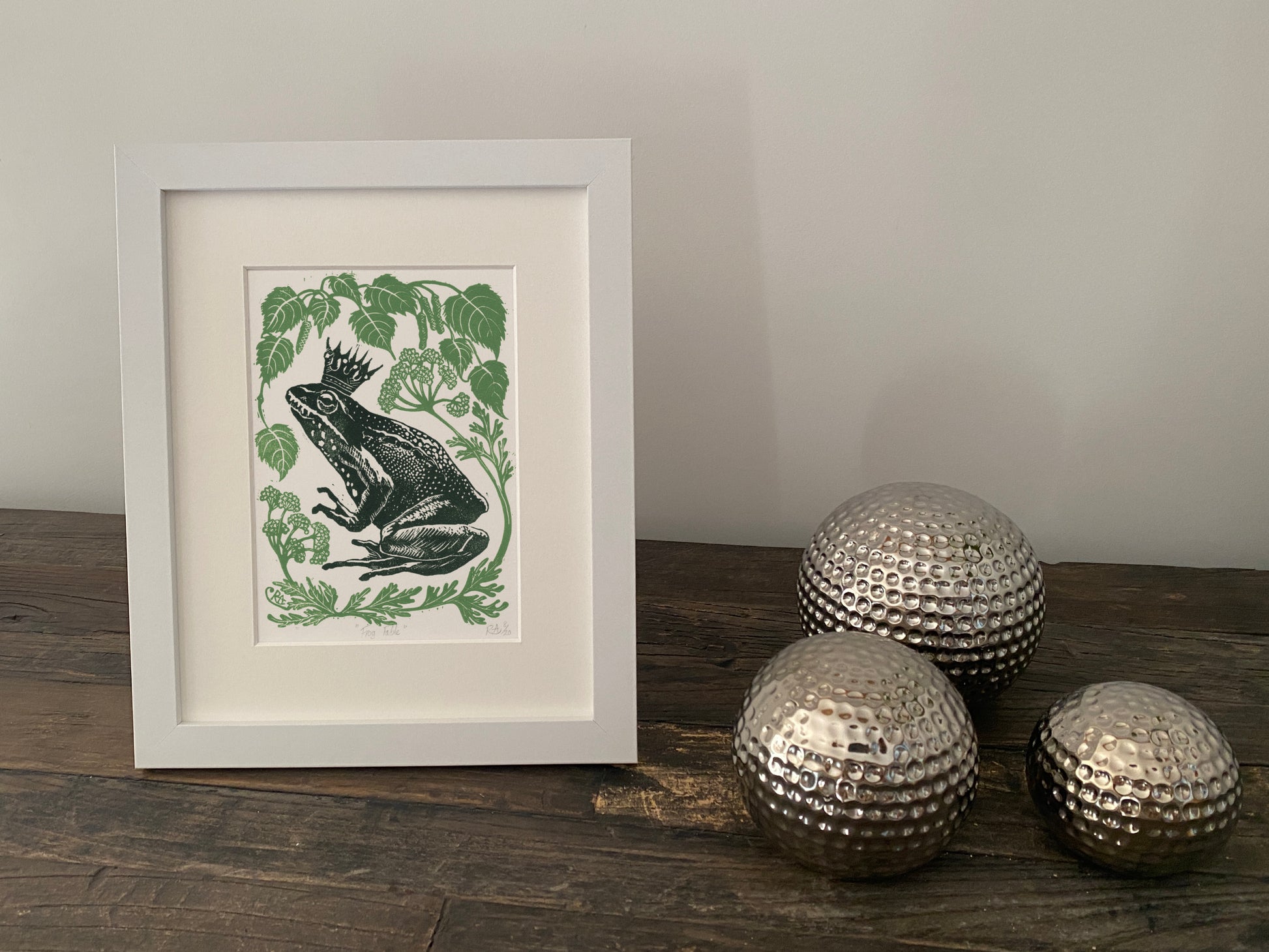 Framed artwork of a frog and leaves on a wooden surface with decorative silver balls.