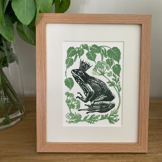 Framed artwork of a frog with a crown surrounded by leaves on a wooden surface.