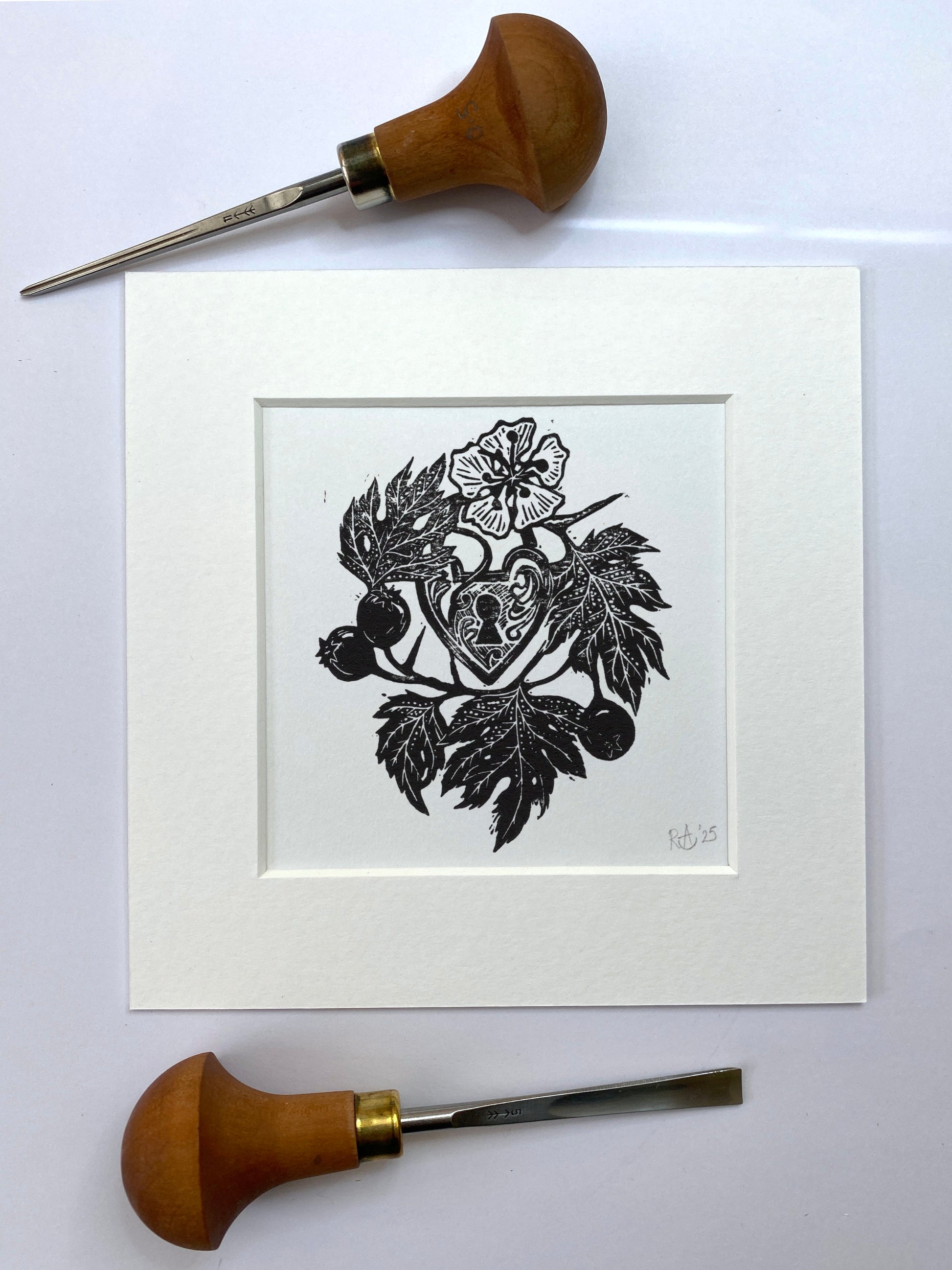 Woodcut print of heart lock surrounded by leaves and flowers, framed with two wood carving tools on a white background.