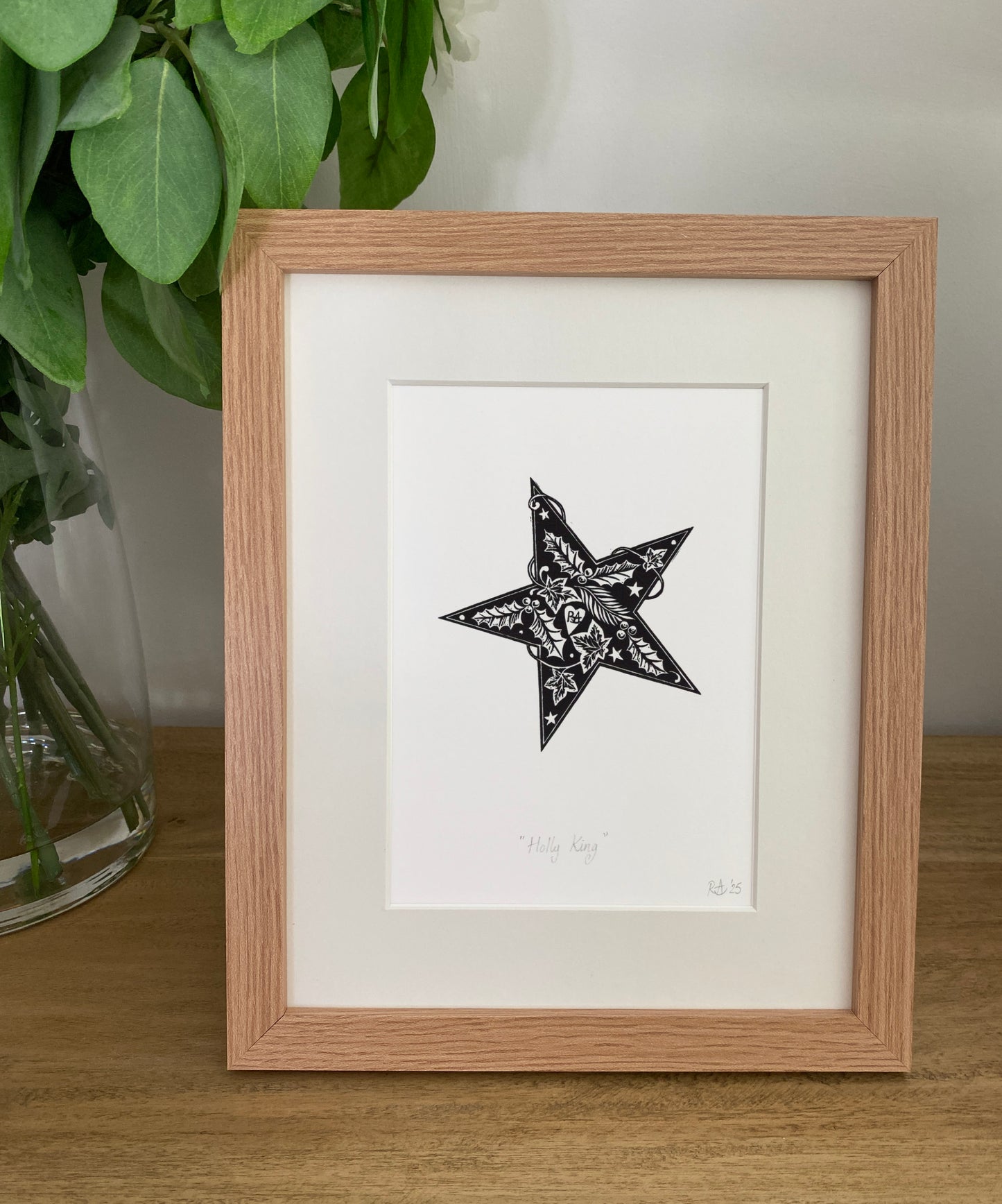 Framed artwork of a black star design on a white background, placed on a wooden surface with greenery in the background.