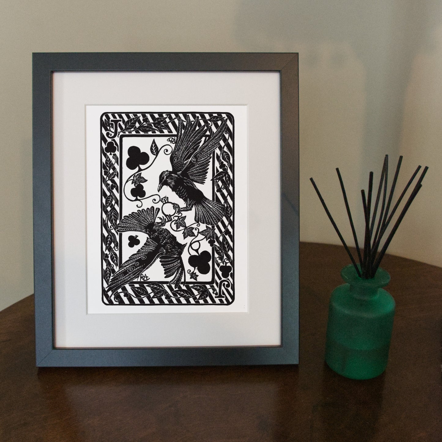 Framed black and white artwork of birds on a white background, placed on a wooden surface with a green vase.