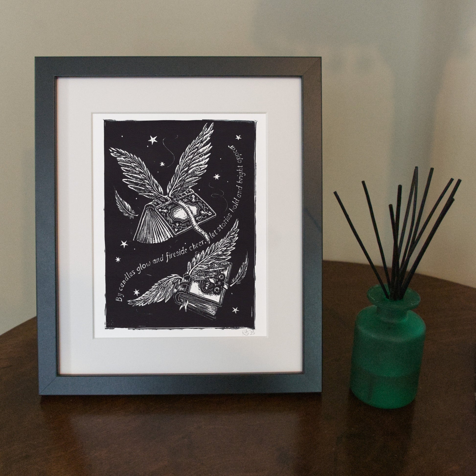 Framed black and white artwork of a winged creature with a book on a wooden surface next to a green vase with sticks.