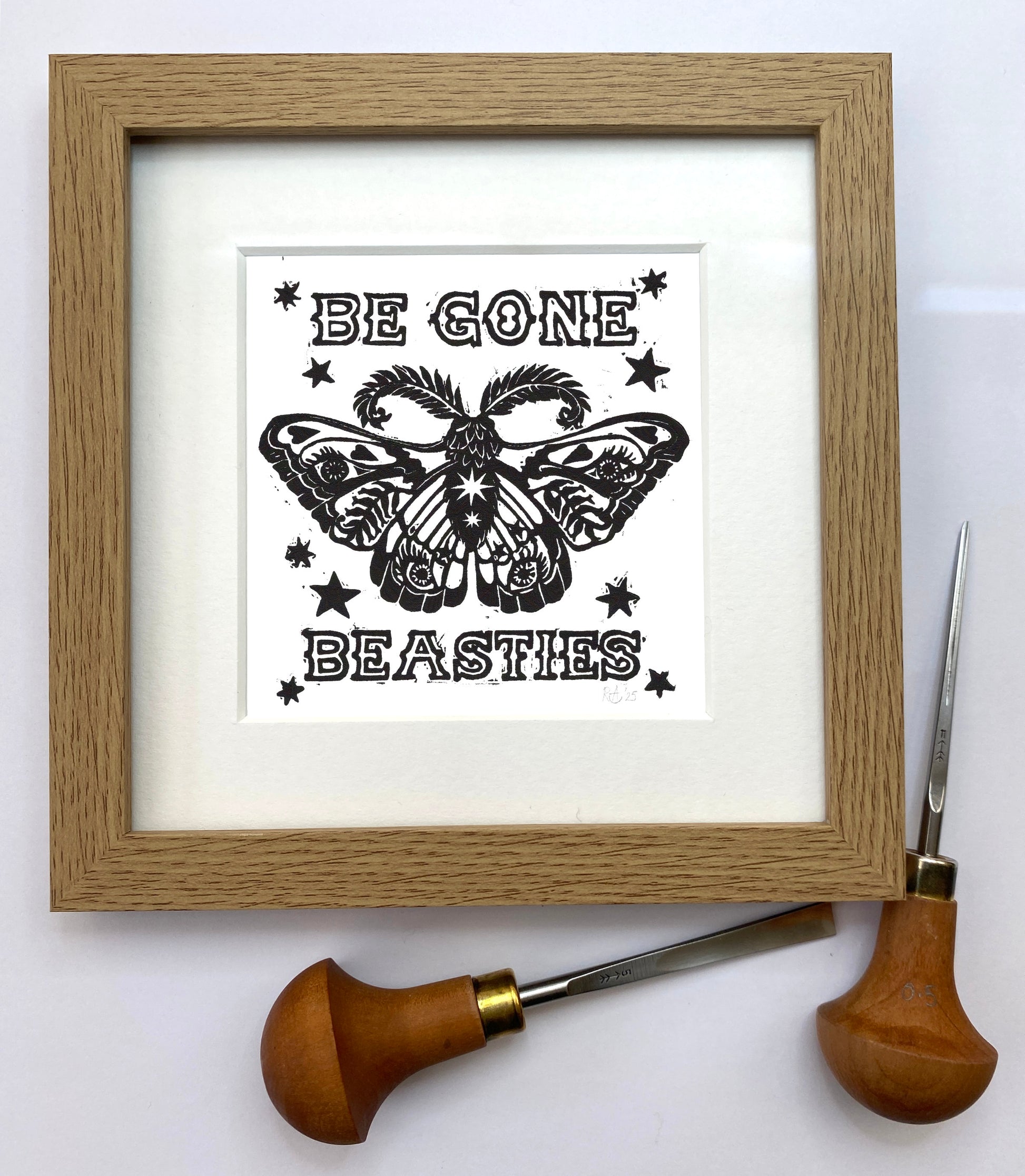 Framed artwork with a butterfly design and text 'Be Gone Beasties' on a white background, with a woodcarving tool beside it.