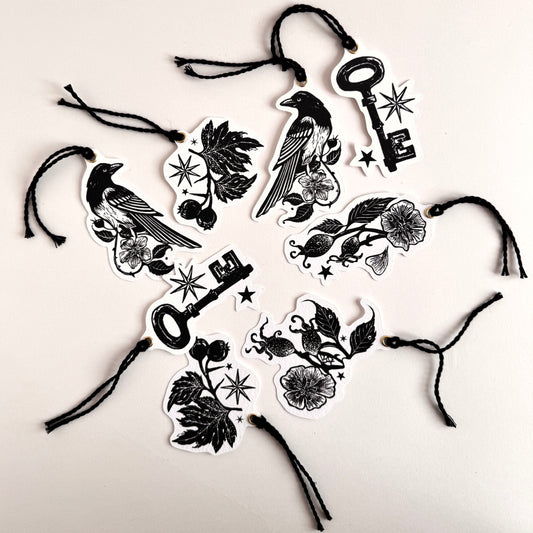 A set of white die-cut stickers featuring black vintage-style illustrations of birds, keys, stars, and botanical motifs, each with a short black hanging cord, arranged on a plain light background.
