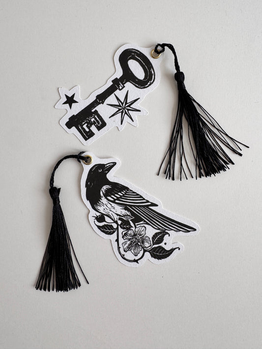 Two black and white paper cutout bookmarks shaped like a key and a bird with floral details, each attached to a black tassel, displayed on a light gray background.