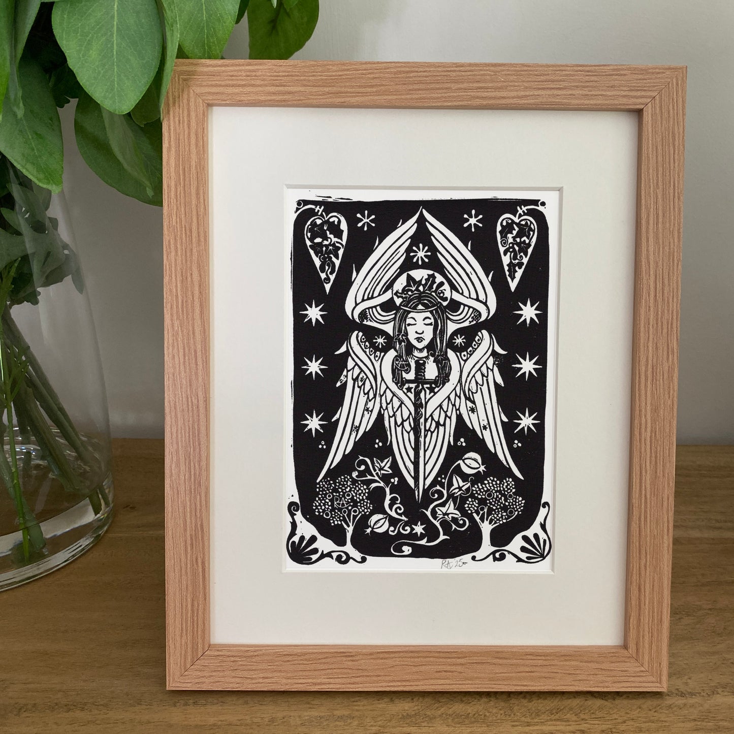 Framed black and white artwork of an angel on a wooden surface with a plant in the background.
