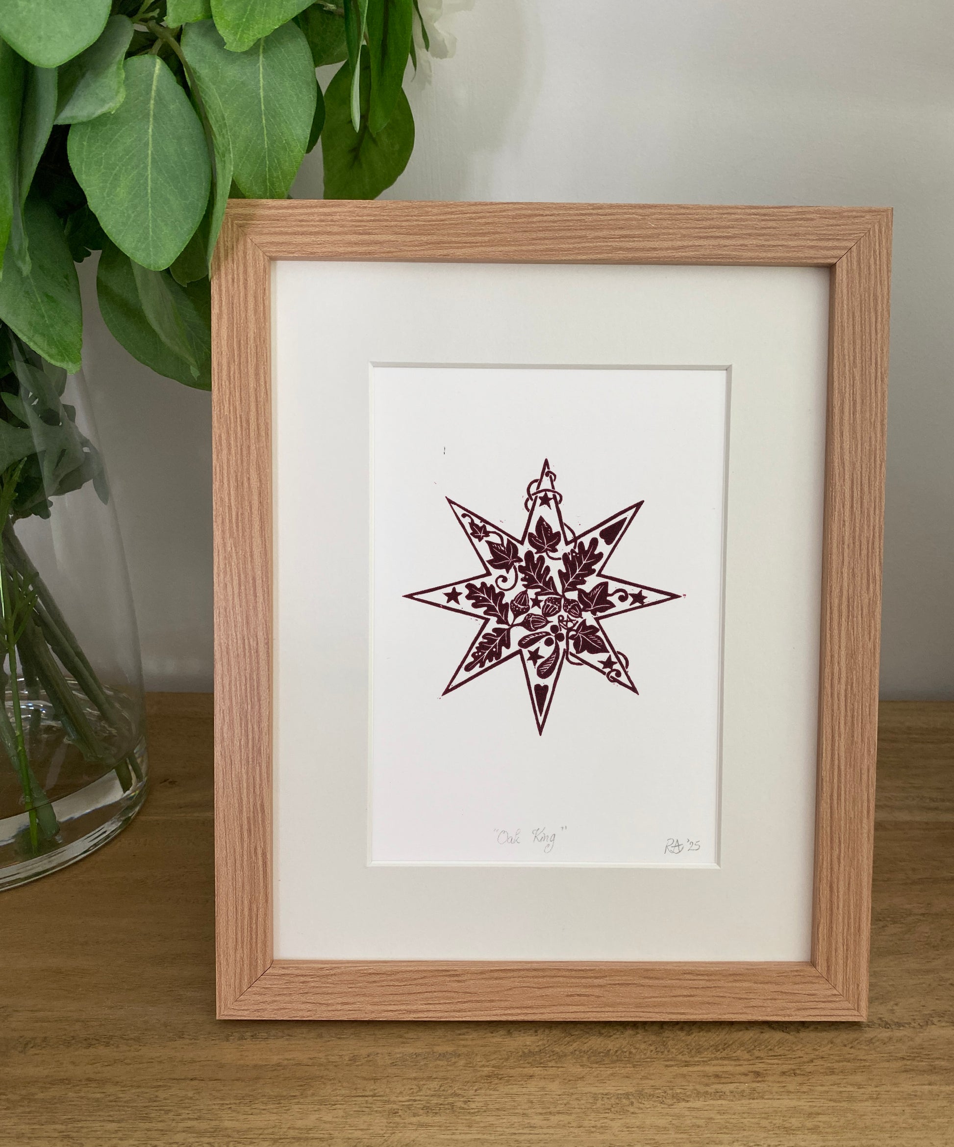 Framed artwork of a star design on a white background, placed on a wooden surface with green leaves in the background.