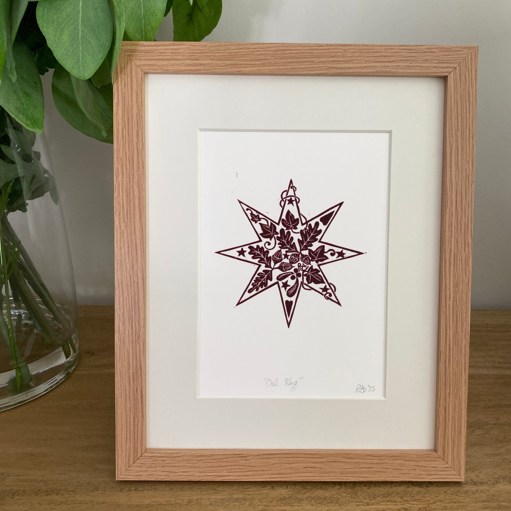 Framed artwork of a star design on a white background, placed on a wooden surface with a plant in the background.