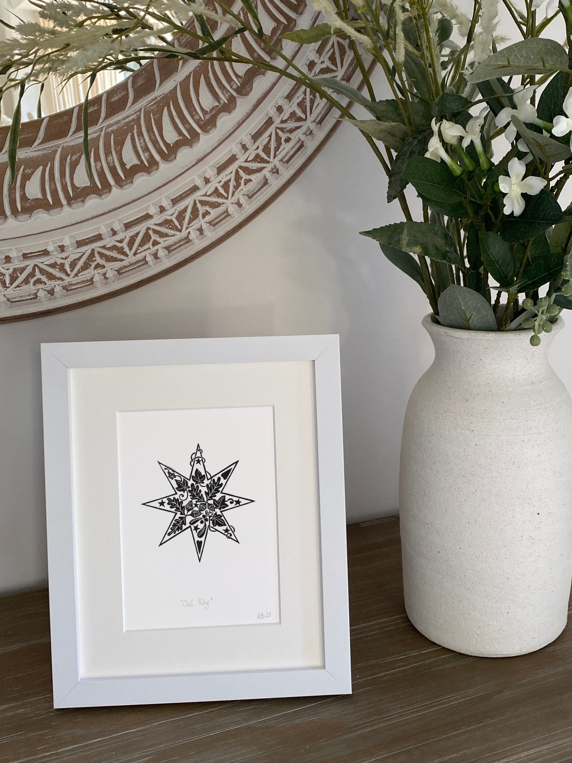 Framed black and white artwork on a wooden surface with a vase of flowers and decorative mirror in the background.