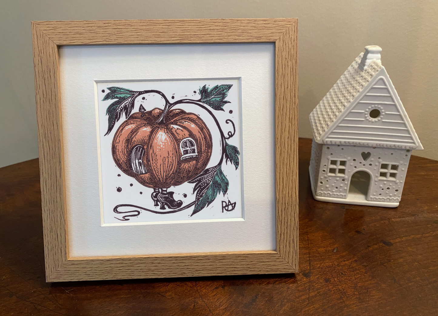 Framed illustration of a pumpkin with a house design on a wooden surface next to a small white house ornament.