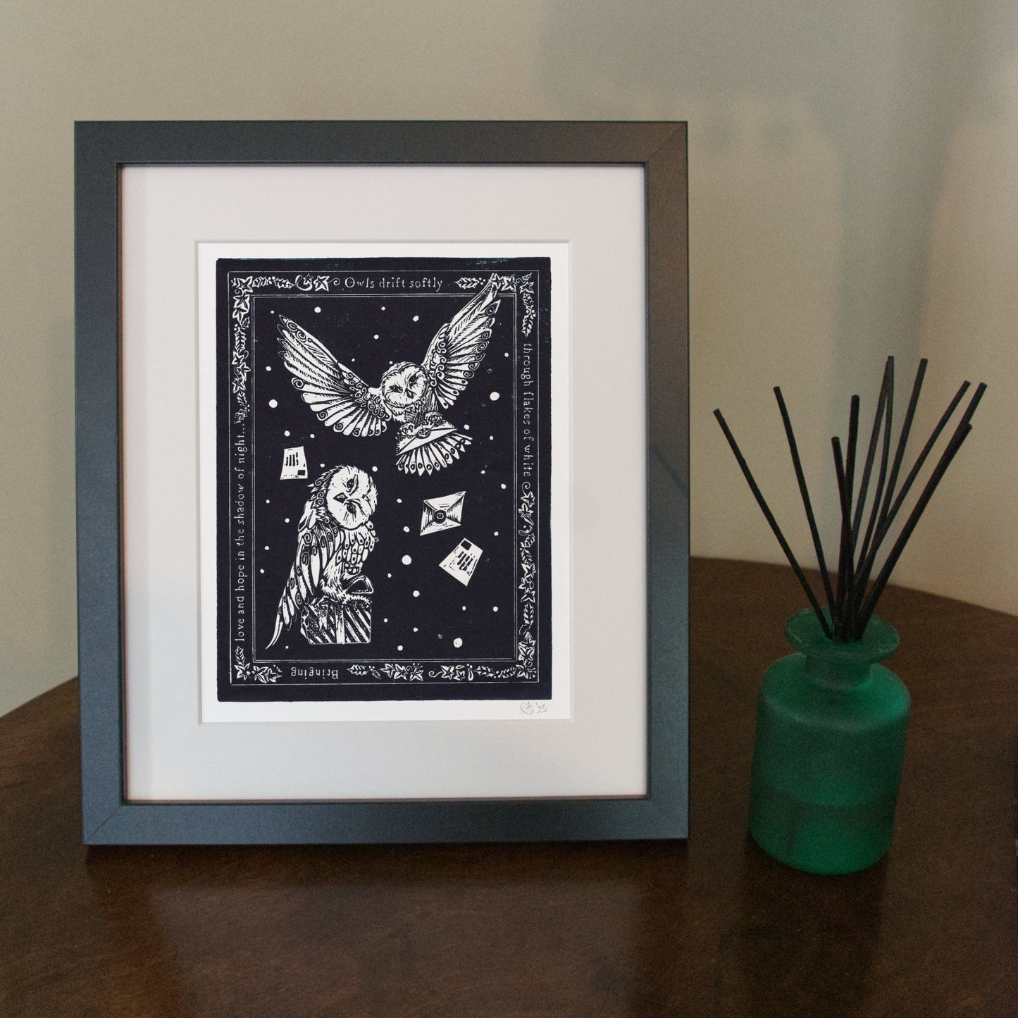 Framed artwork of an owl with decorative elements on a wooden surface next to a green vase with black sticks.