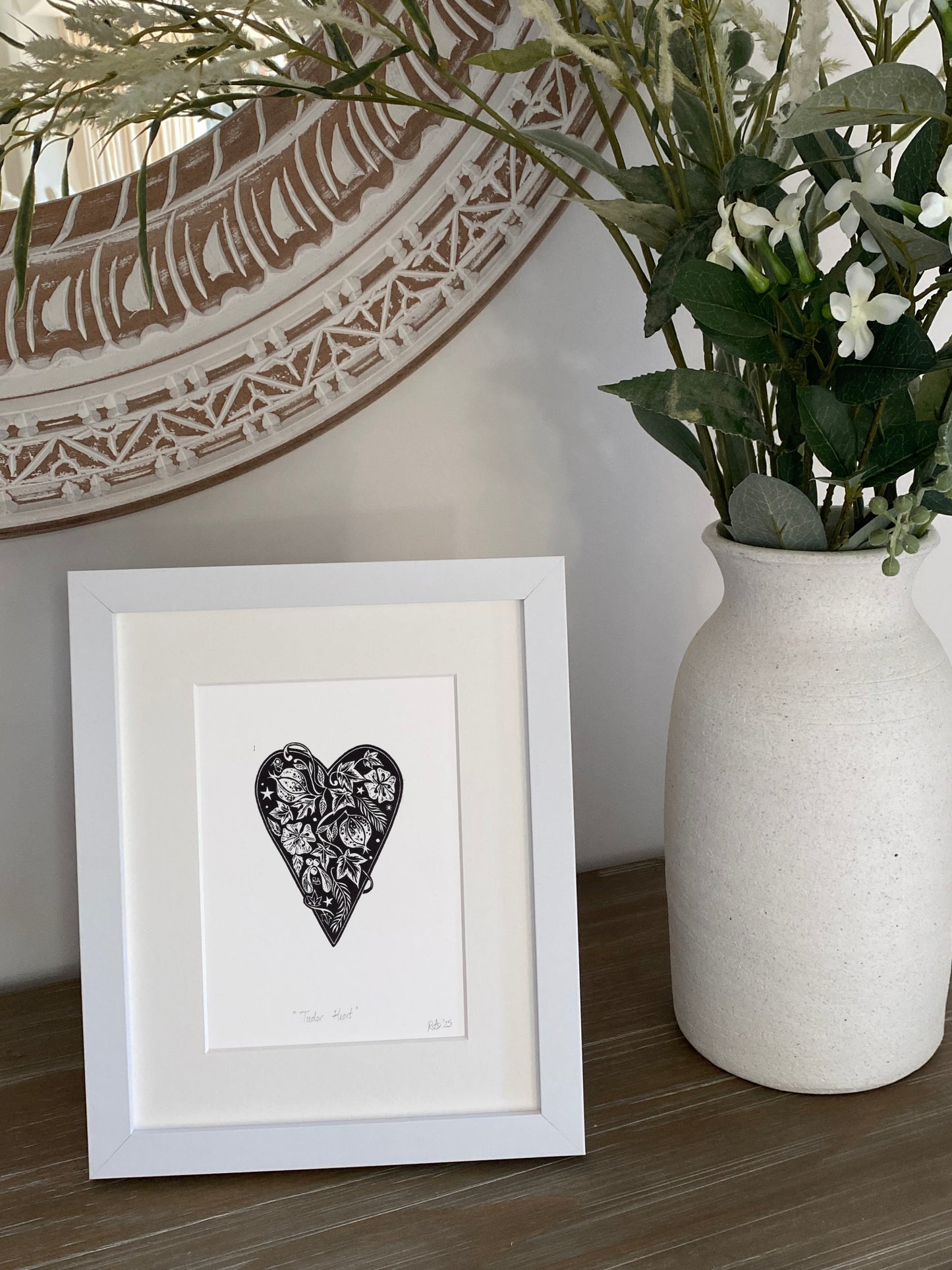 Framed artwork with a heart design next to a vase with plants on a wooden surface.