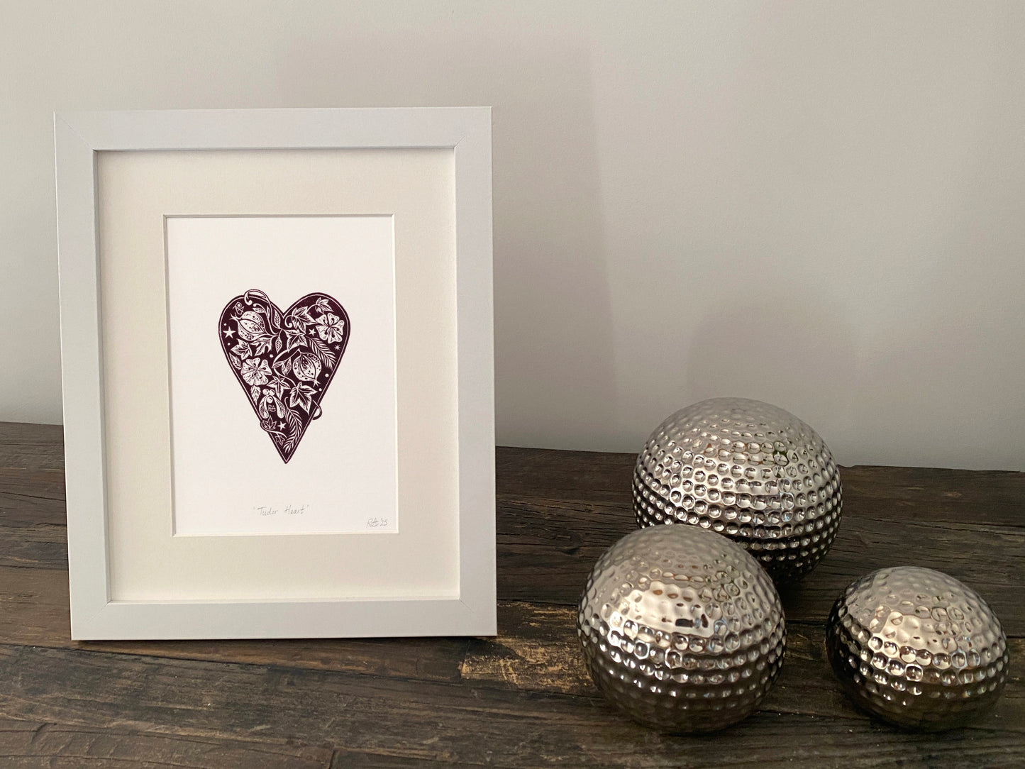 Framed artwork of a heart with decorative elements on a wooden surface with metallic spherical objects.
