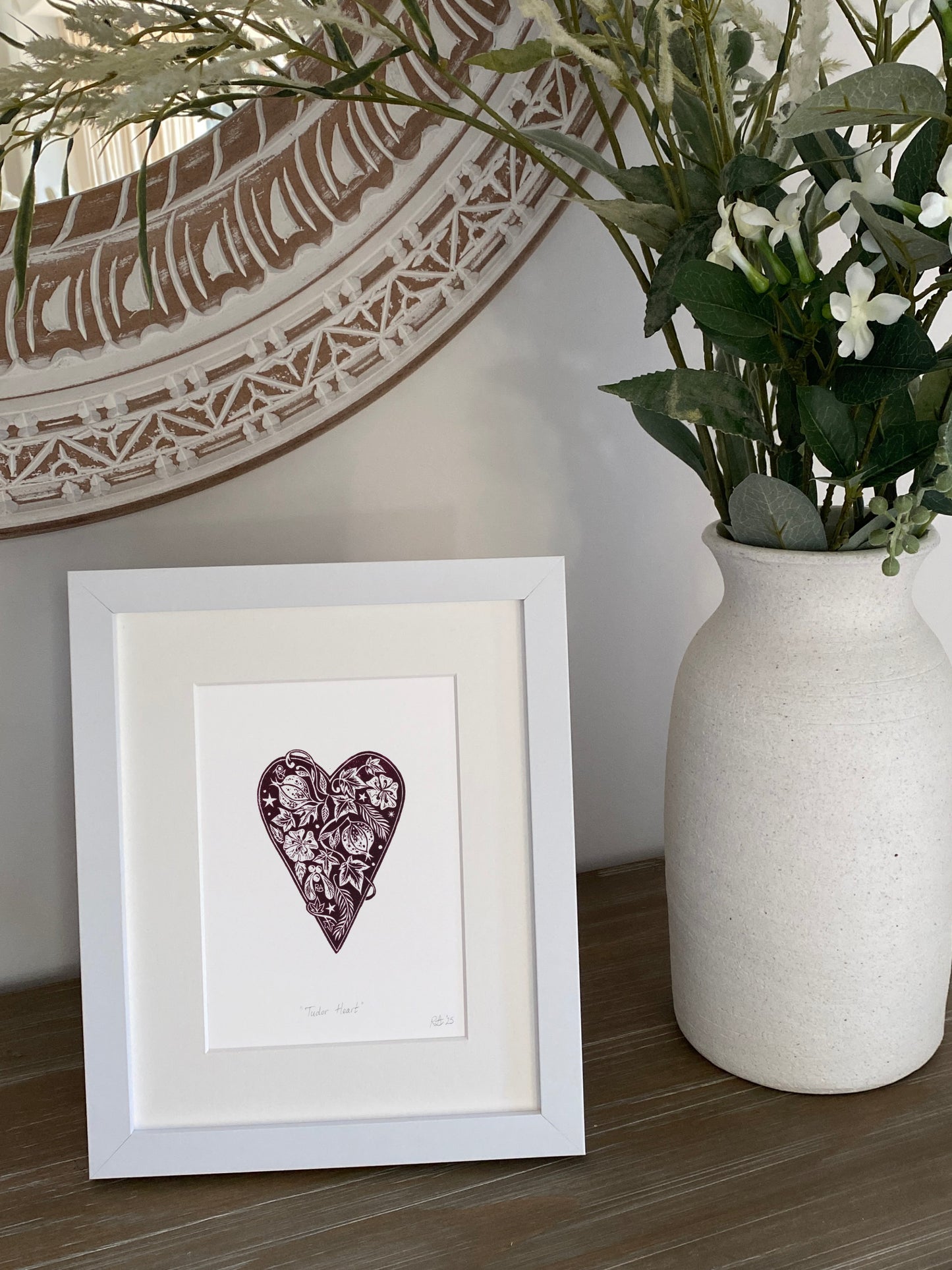 Framed artwork with a heart design next to a vase with plants on a wooden surface.