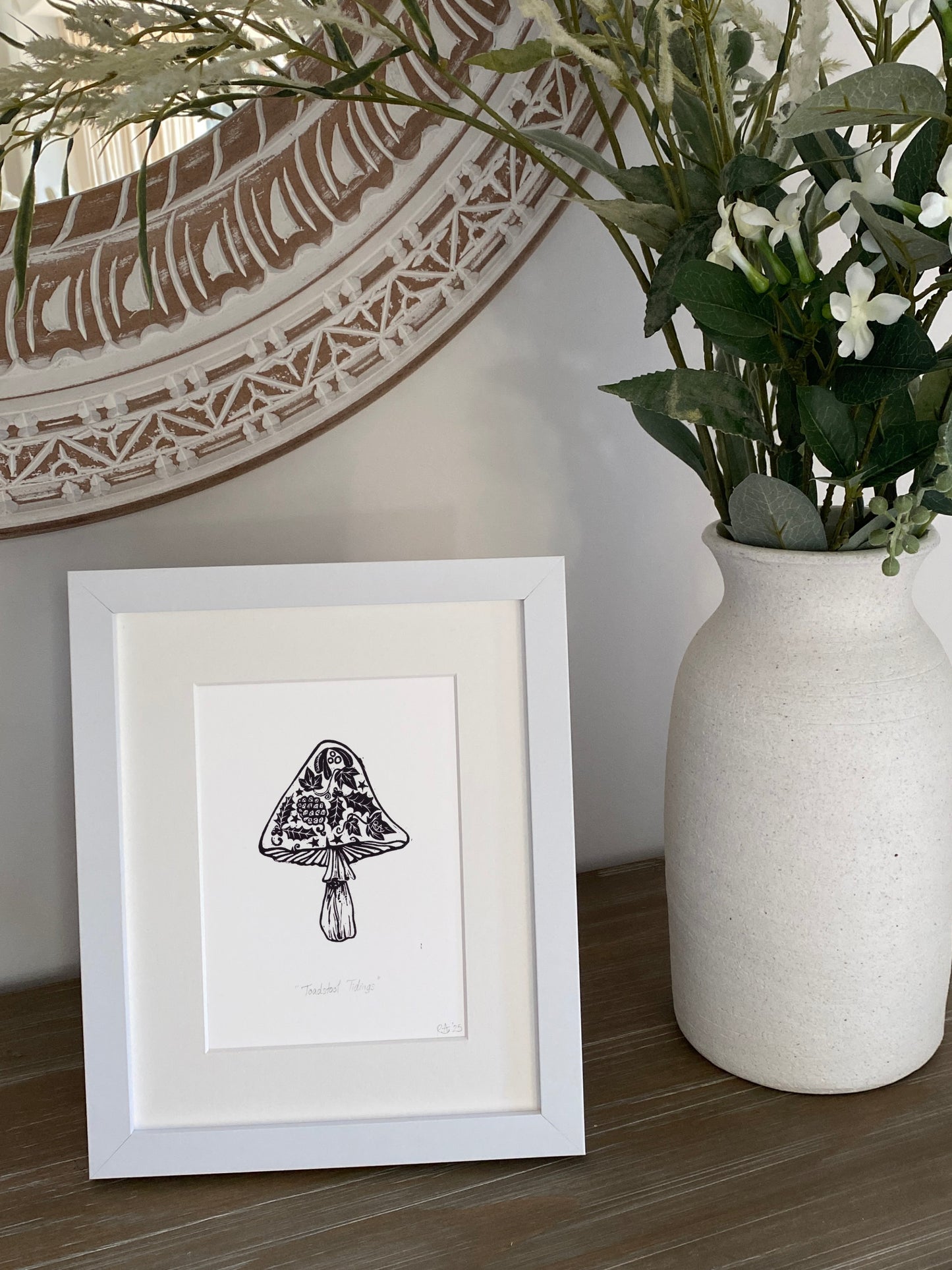 Framed artwork of a mushroom next to a vase with plants on a wooden surface.