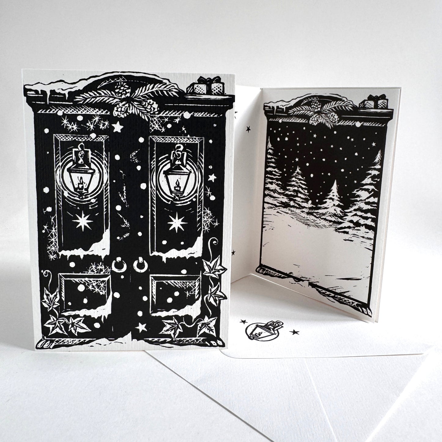 A black and white illustrated greeting card set shown on a white background, with one card standing flat and another opened to show a snowy night scene. The card design features lanterns, stars, snowflakes, and decorative border details.